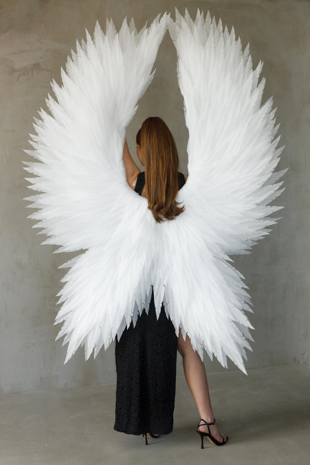 May include: A person wearing large, white feather angel wings, with a black dress. The wings are spread wide, creating a dramatic effect. The background is a plain, light gray wall.