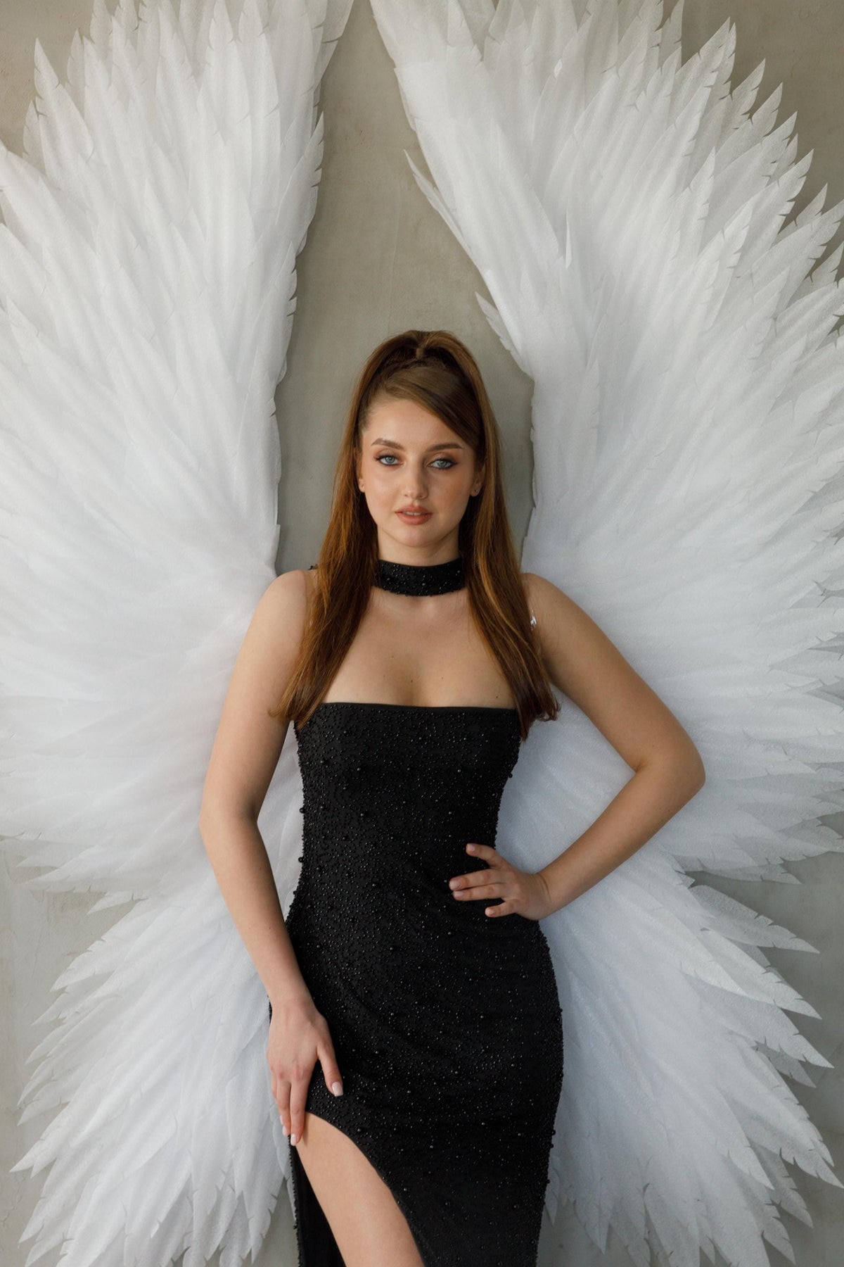 May include: A woman wearing a black beaded strapless dress with a high slit, a black choker, and large white feathered angel wings. The wings are spread wide, creating a dramatic effect. The background is a neutral gray.