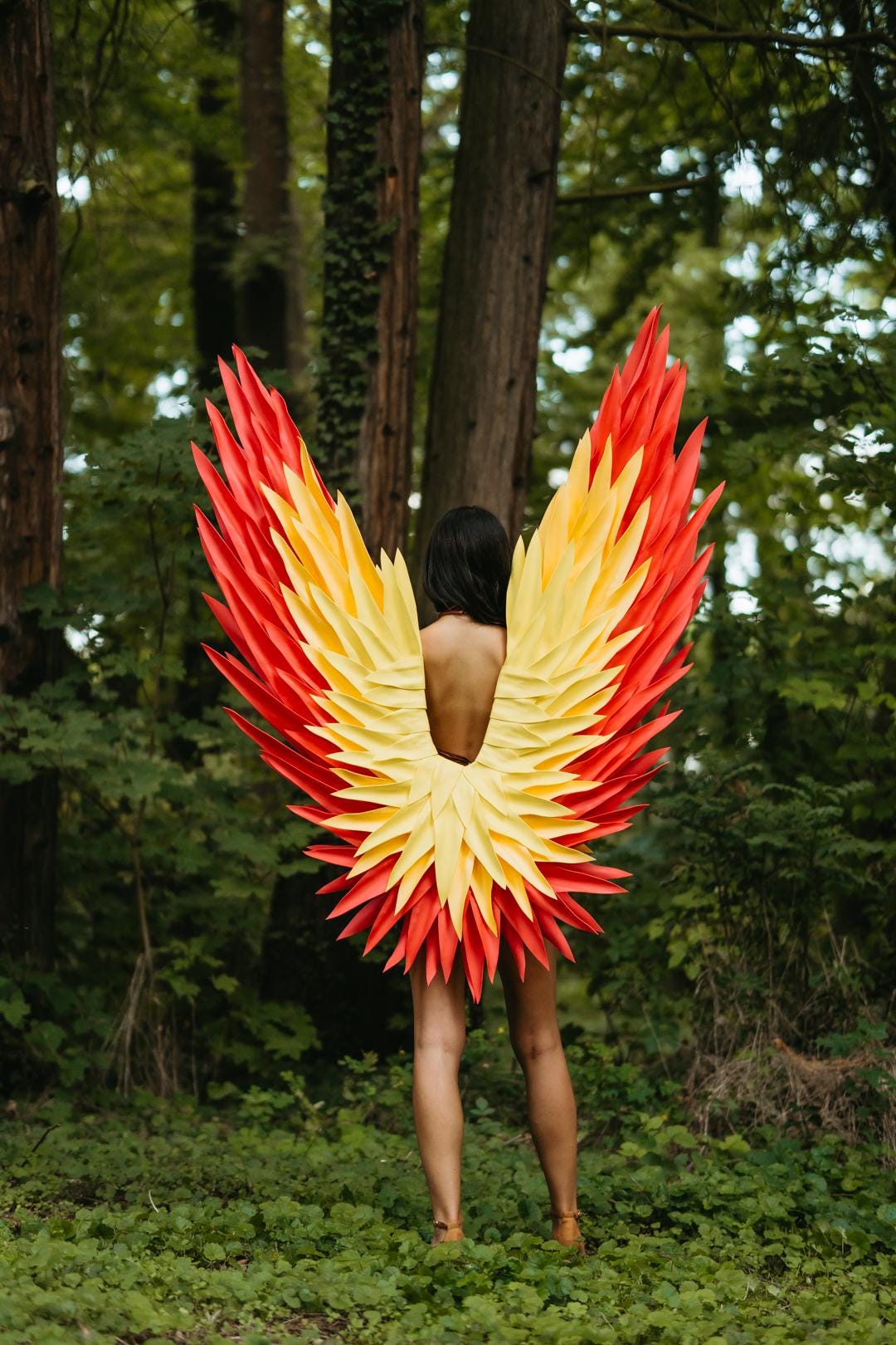 May include: A pair of large, wearable wings featuring a layered feather design in a vibrant gradient of bright yellow and deep red. The wings are constructed with pointed, individual feather-like segments that fan out from the center to create a full, expansive shape.