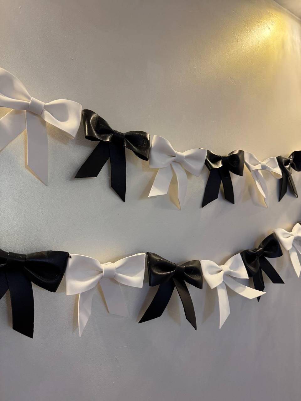 May include: A decorative garland of alternating black and white bows. The bows are made of a smooth material and are arranged in two rows against a neutral wall. The bows are tied in a classic bow shape.