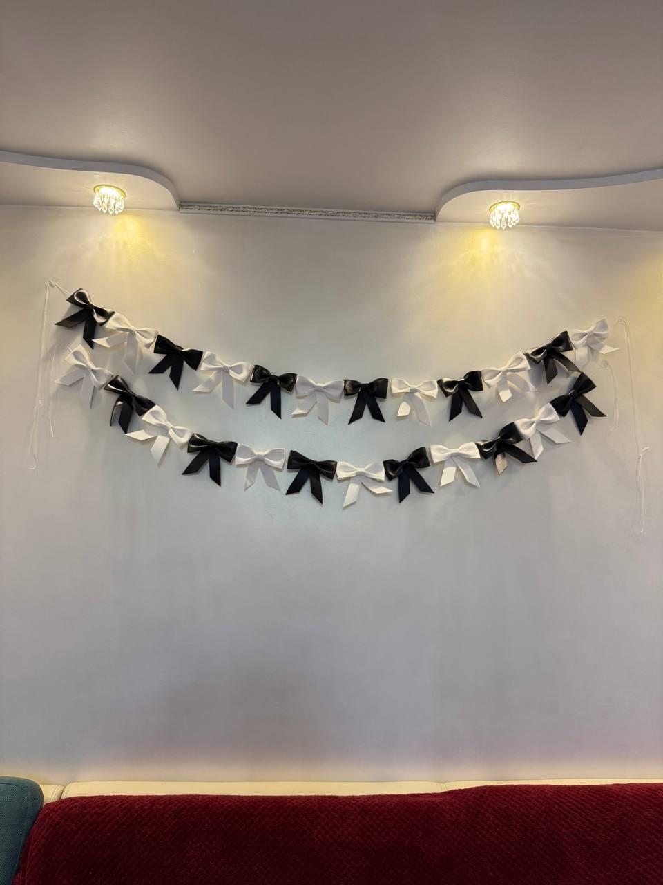 May include: Two strands of decorative bows, alternating between black and white, hang against a white wall. The bows are evenly spaced and appear to be made of a satin-like material. The overall aesthetic is simple and elegant.