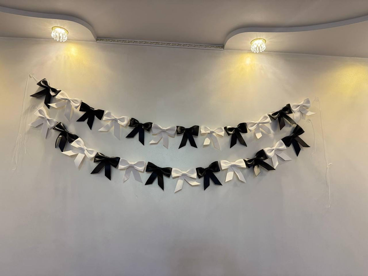 May include: A decorative garland of alternating black and white bows hangs against a white wall. The bows are evenly spaced and arranged in two parallel rows, creating a festive and elegant look. Two small, round lights are in the background.