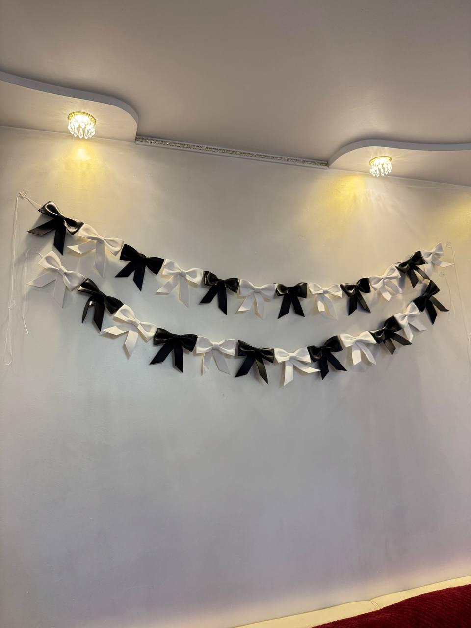 May include: A decorative garland of alternating black and white bows hangs against a white wall. The bows are evenly spaced and strung together, creating a festive and elegant look. Two small, decorative lights are mounted on the ceiling above the garland.