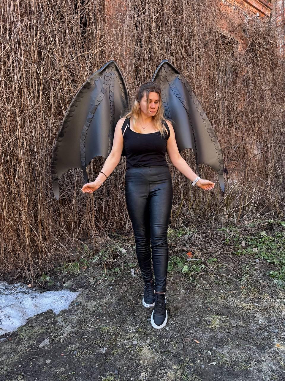 May include: A pair of large, black, articulated dragon wings with a textured, scale-like pattern along the edges, worn over a black tank top, black fitted trousers, and black sneakers with white soles, creating a dark, fantasy-inspired costume.