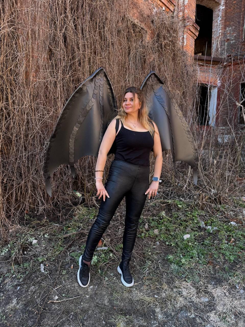 May include: A pair of large, dark gray dragon wings with a textured, scale-like pattern along the edges. The wings are worn as a backpack accessory, featuring a structured frame and pointed tips, paired with a black tank top and black leggings.