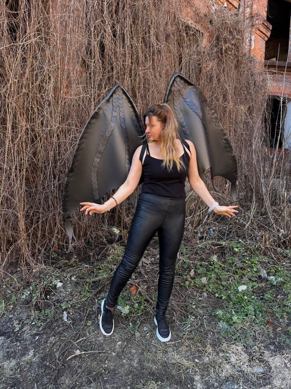 May include: Large black dragon-style wings featuring a layered scale texture and pointed tips. The wings are attached to a black tank top, paired with black fitted trousers and black sneakers. The design mimics a mythical creature's anatomy.