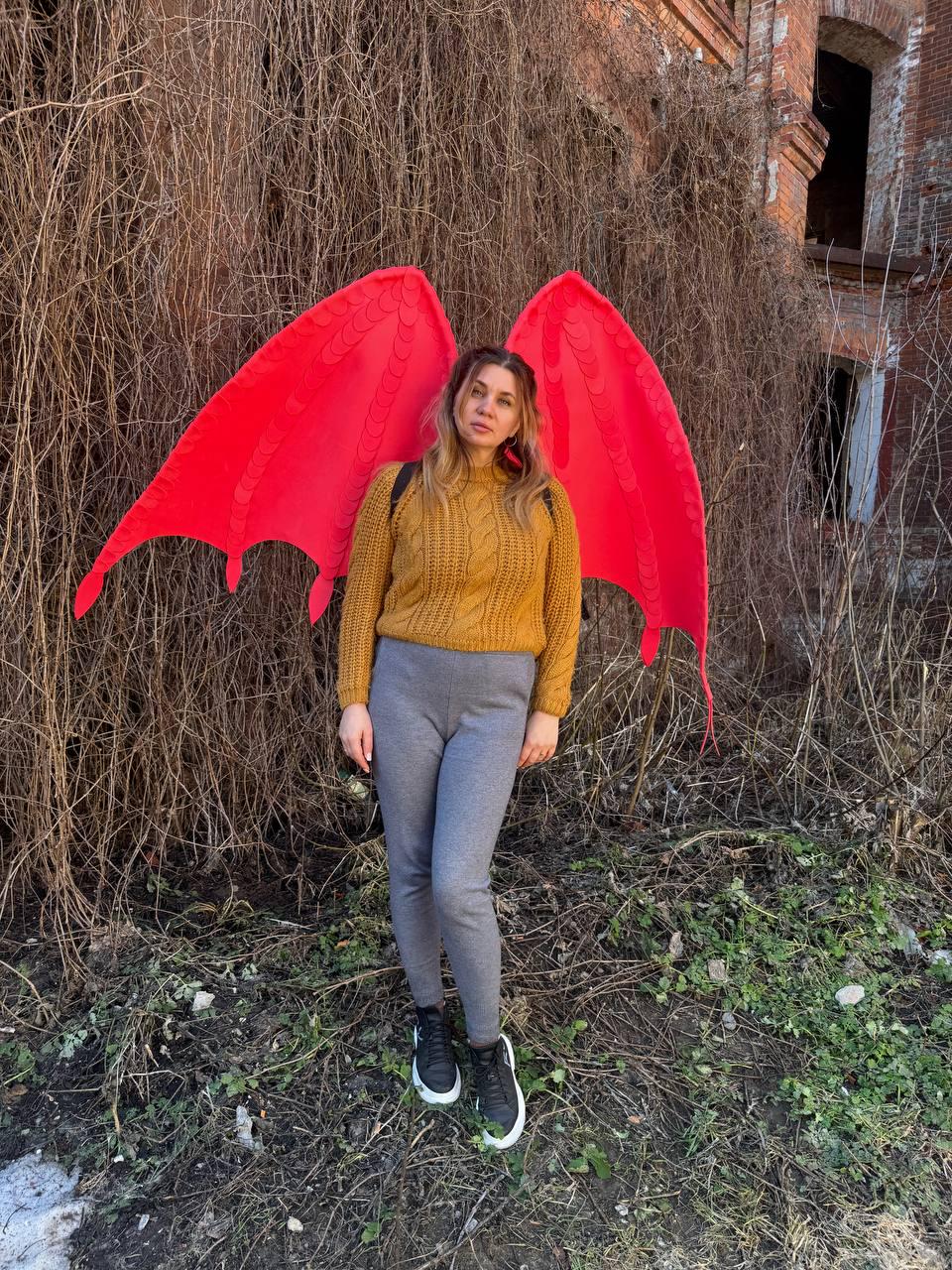 May include: A person wearing a mustard yellow cable knit sweater, gray leggings, and black sneakers with white soles. Large, bright red dragon wings are attached to the person's back. The background features overgrown vines and a weathered brick building.