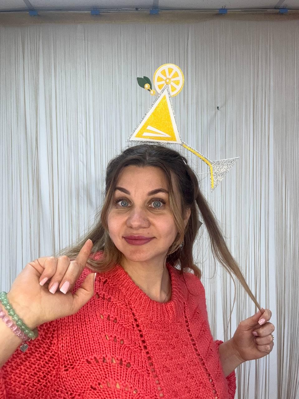 May include: A woman wearing a coral-colored sweater and a cocktail-themed headpiece. The headpiece features a yellow triangle and a lemon slice, with a silver stem. The woman is pointing her index finger.