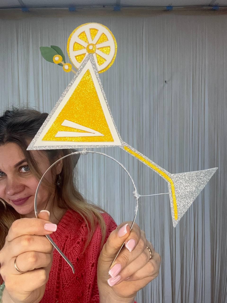 May include: A whimsical headband featuring a glittery yellow martini glass design with a lemon slice and leaves. The cocktail glass is outlined in silver glitter and has a yellow stem. A fun accessory for parties or themed events.