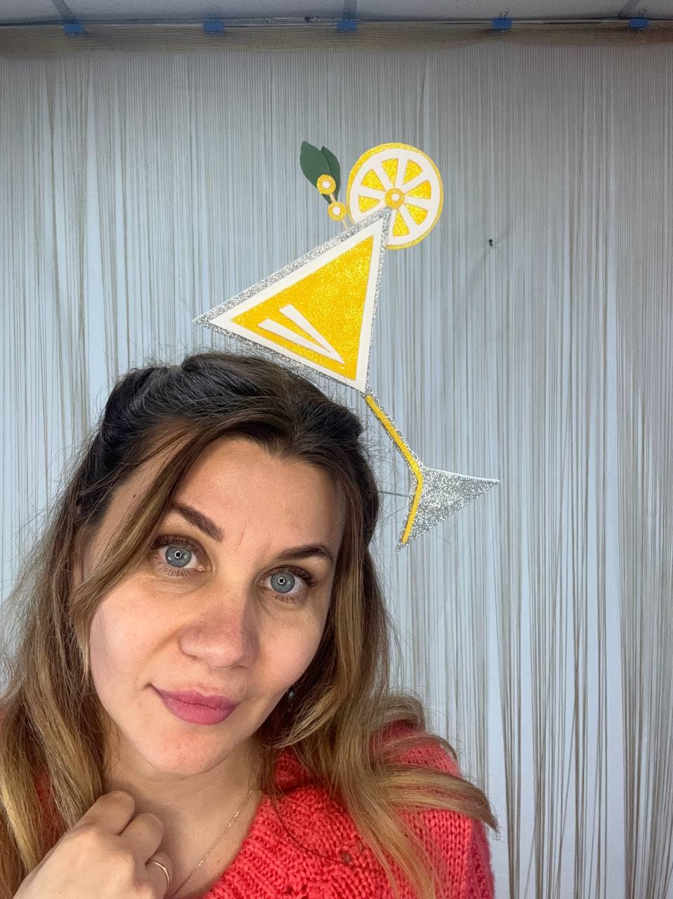 May include: A yellow and silver cocktail-themed headpiece. The headpiece features a yellow triangle, a lemon slice, and a silver stem. The design is made of glittery material and is worn on the head. The background is a light gray curtain.