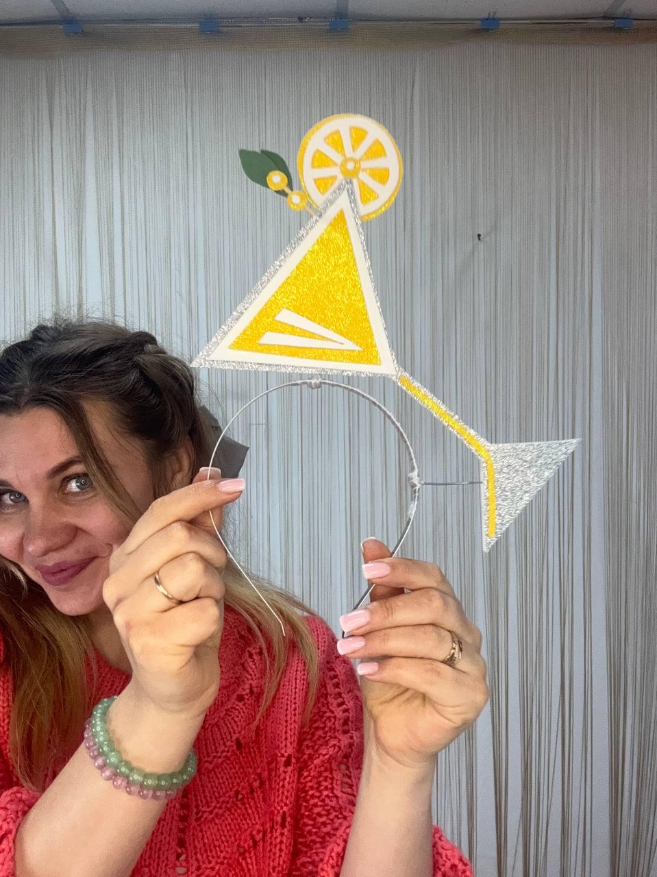 May include: A whimsical headband featuring a yellow and silver glitter martini glass design. The cocktail-themed accessory includes a lemon slice and a green leaf accent. The headband is made of silver metal and is perfect for parties or themed events.