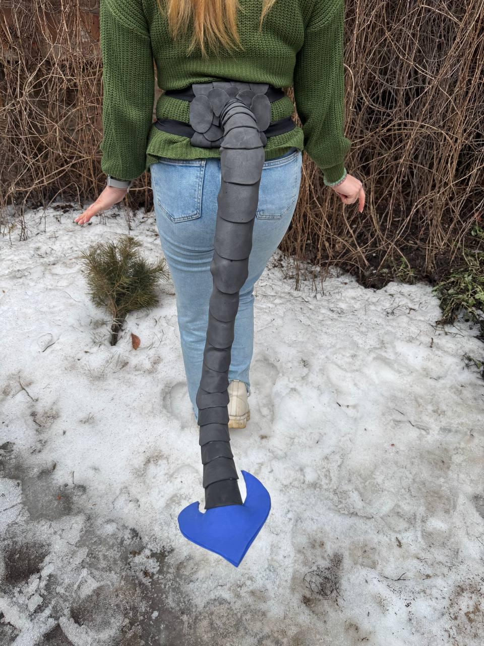 May include: A black and blue costume tail, attached to a black belt. The tail is segmented and tapers to a blue, heart-shaped tip. The costume is worn over blue jeans and a green sweater.