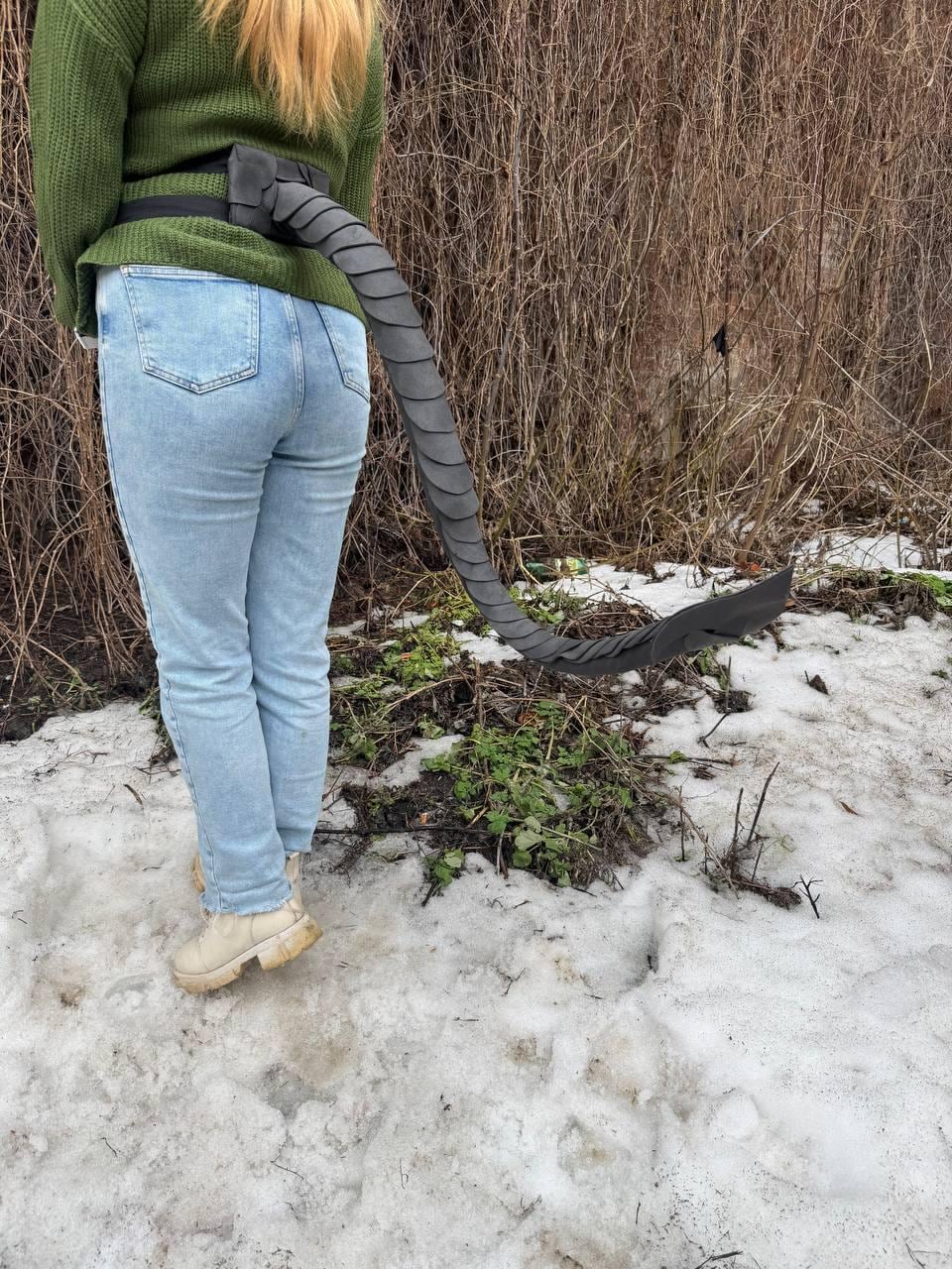 May include: A black, segmented tail prop attached to a black belt. The tail is long and curved, with a series of overlapping, scale-like segments. The prop is worn with light blue jeans and a green sweater. The background is a snowy, outdoor scene.