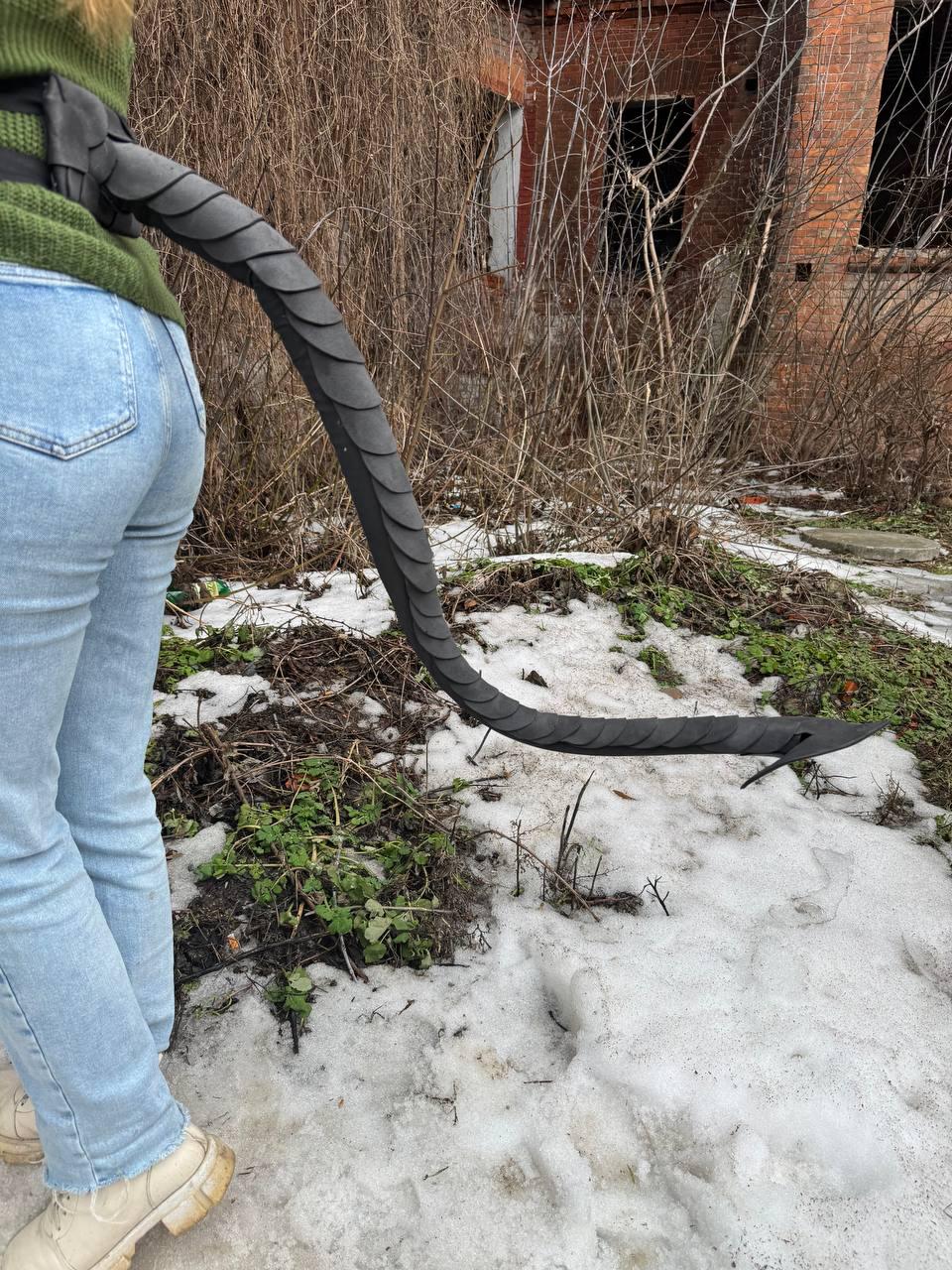 May include: A black, segmented tail prop, possibly for cosplay or costume use, extends from a harness. The tail is made of dark gray material and is curved. The background includes an old brick building and patches of snow.
