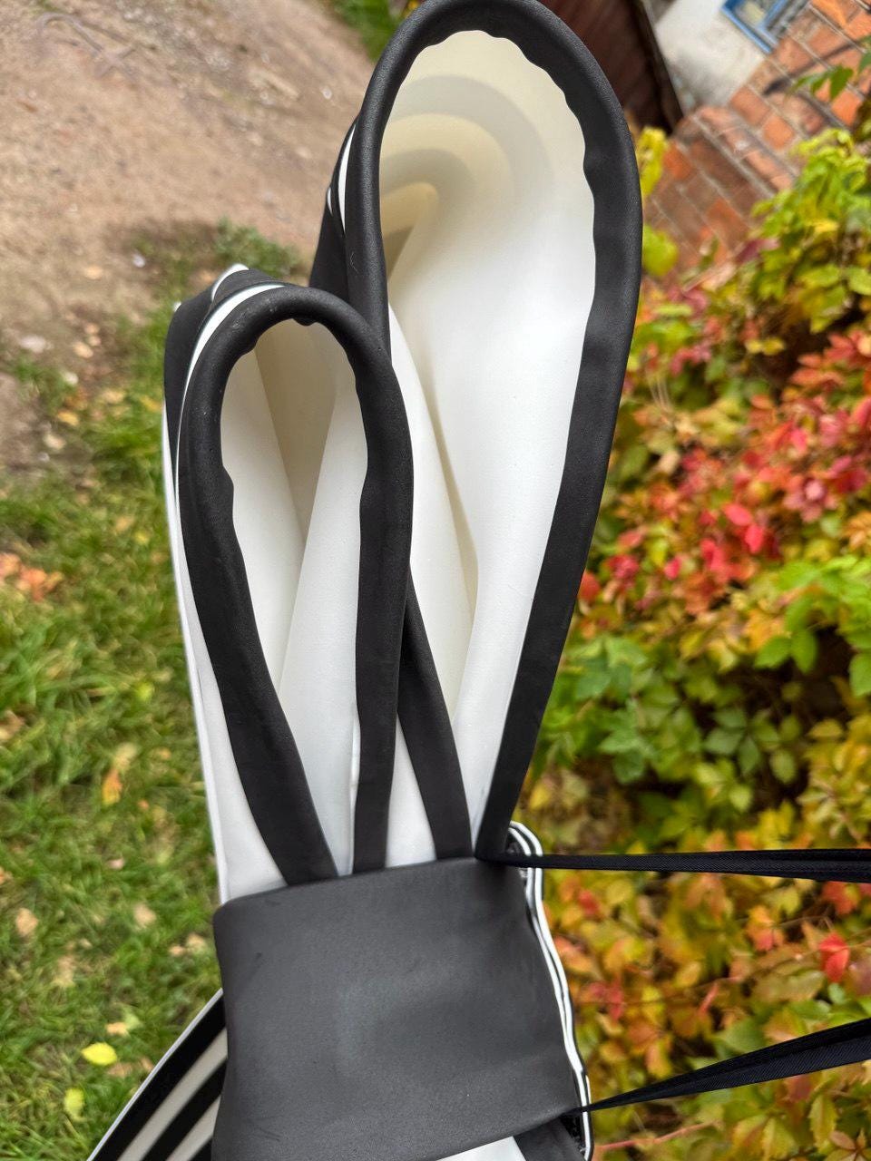 May include: A black and white striped bag with a black handle and trim. The bag has a rectangular shape and is made of a soft, flexible material. The background is blurred, with green grass and colorful foliage.