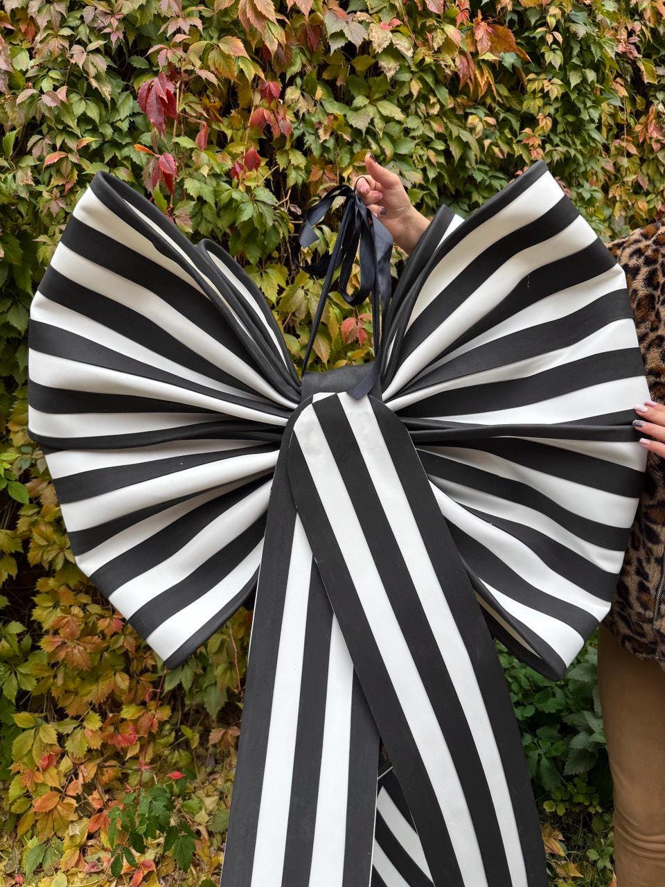 May include: A large, oversized bow with black and white stripes. The bow is held up by a black ribbon. The bow is made of a fabric with a satin-like sheen. The background is a wall of green and red leaves.