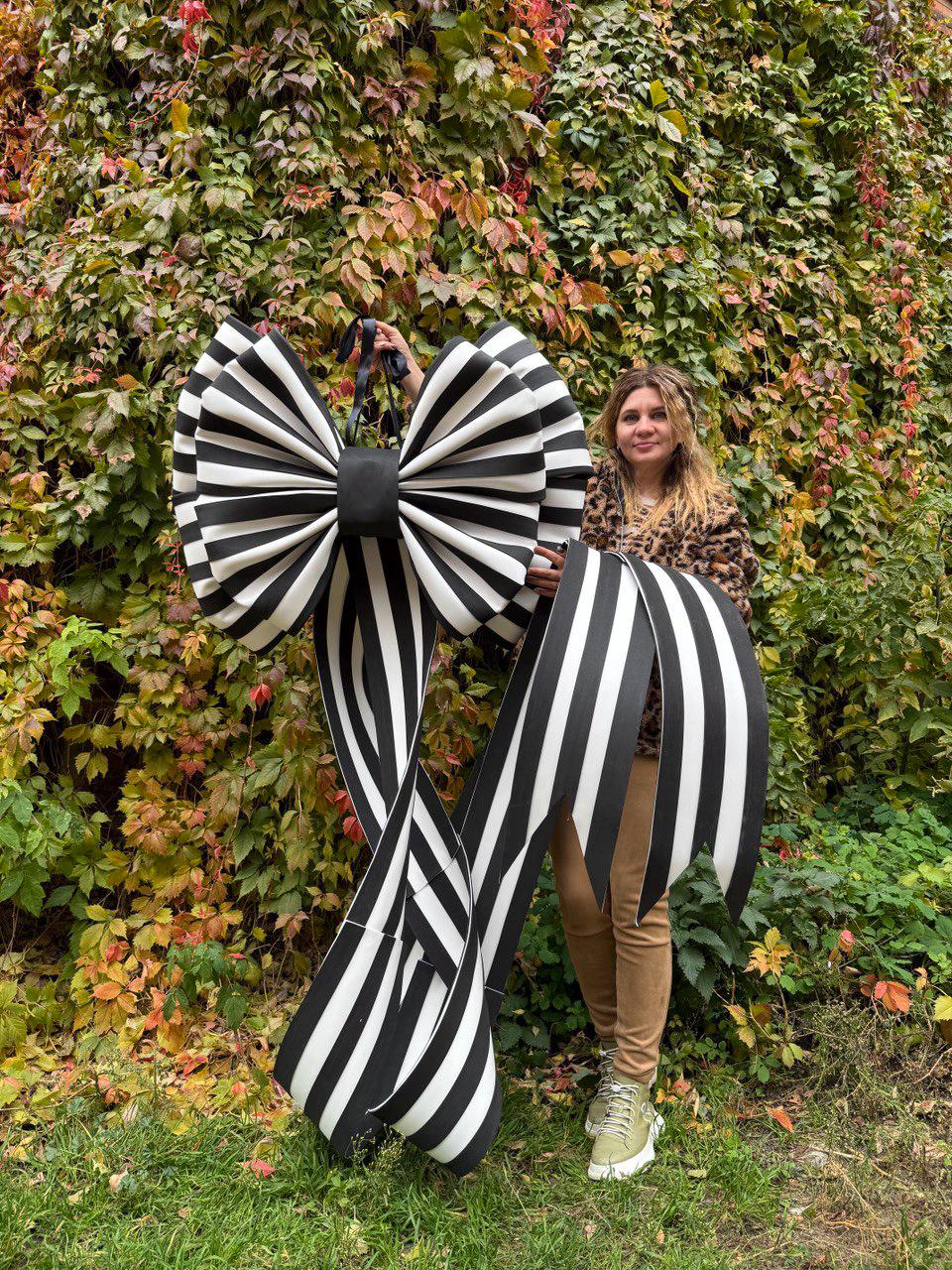 May include: A large, decorative bow with black and white stripes. The bow is held up in front of a background of green and red foliage. The bow has a large center knot and long, flowing tails.