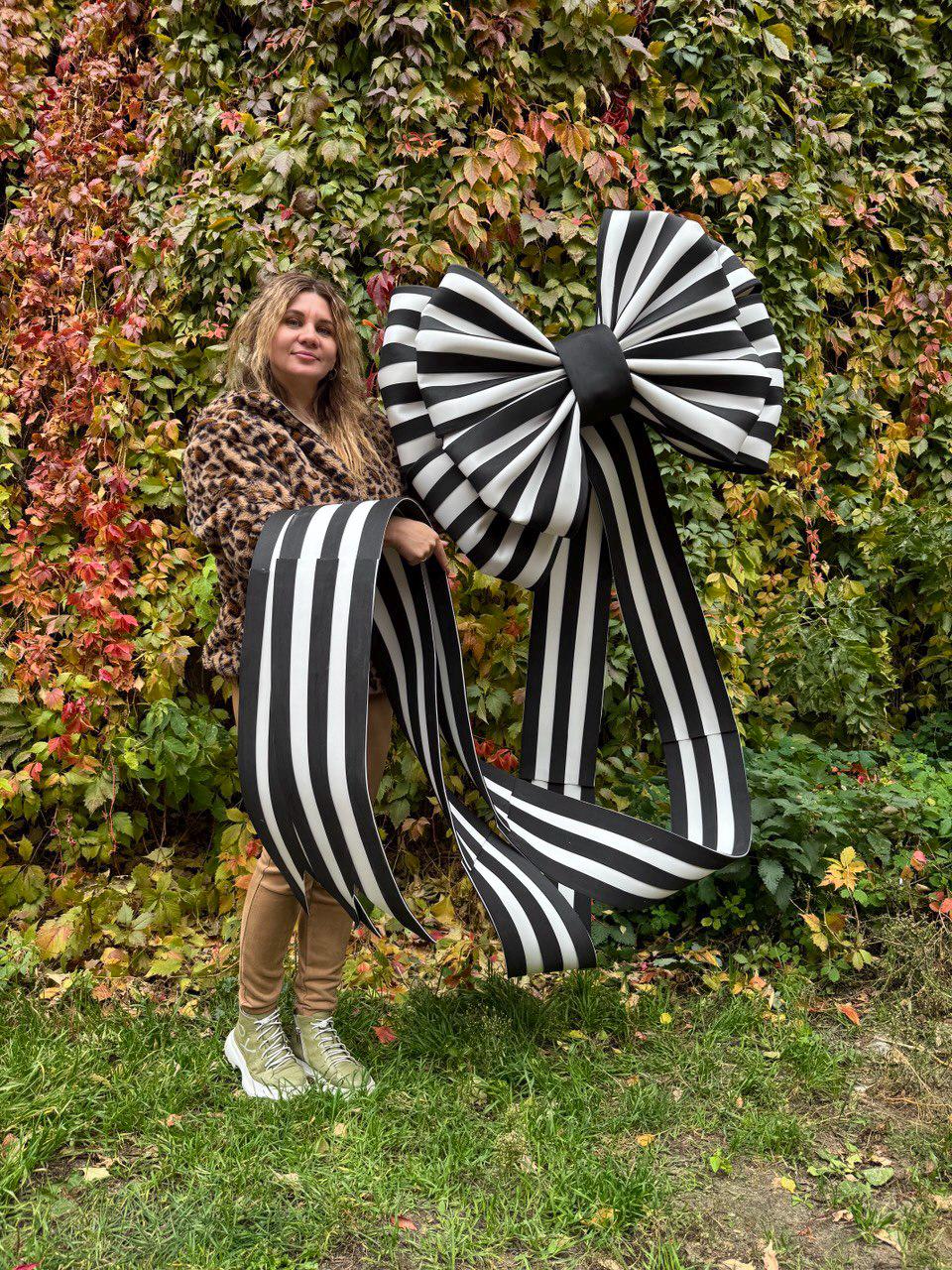 May include: A large black and white striped bow with long, flowing ribbons. The bow is held up in front of a background of green and red foliage. The bow's design features bold, contrasting stripes.