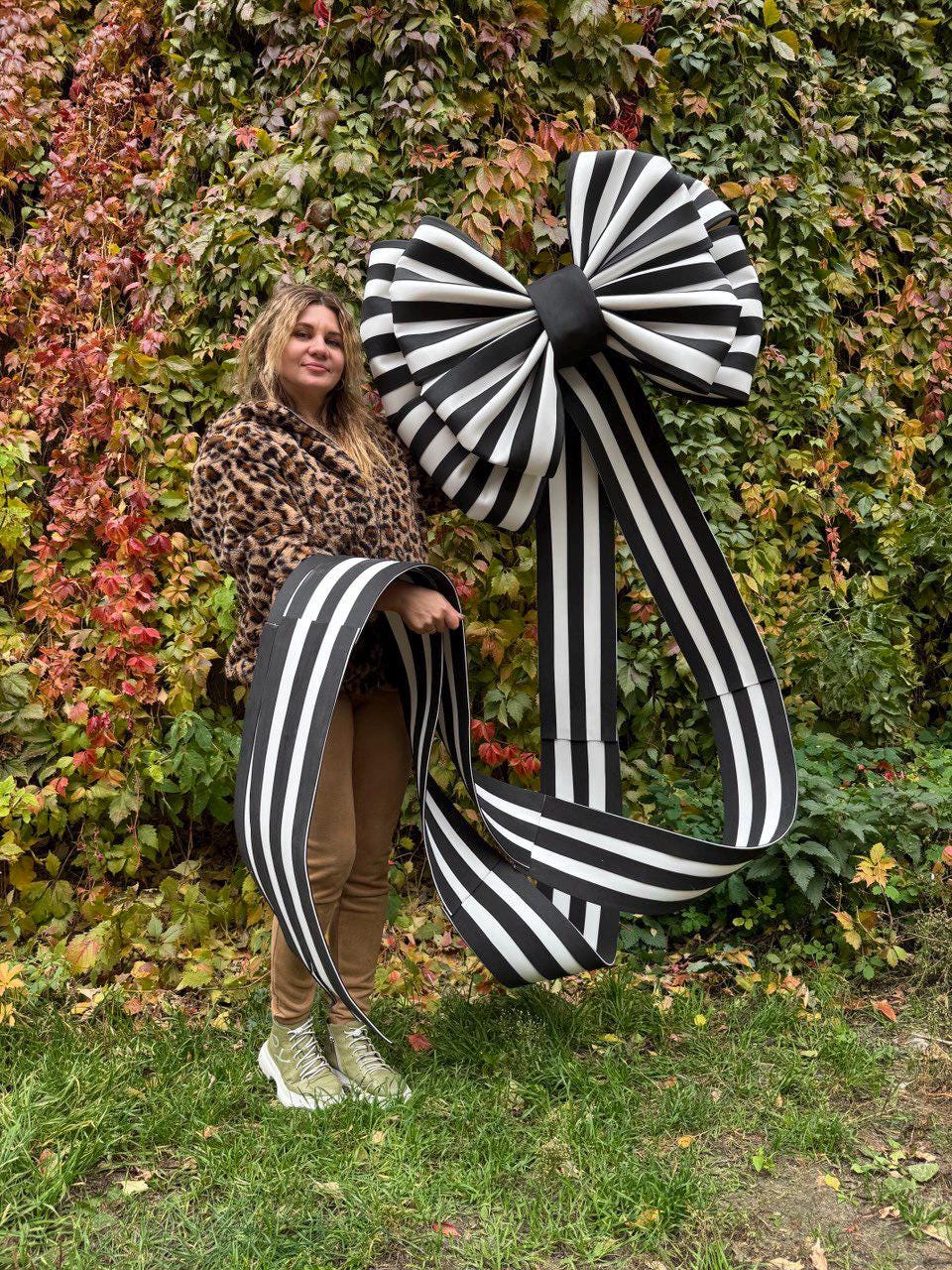 May include: A large black and white striped bow with long, matching ribbons. The bow is held in front of a background of green and red foliage. The bow's design features alternating black and white stripes.