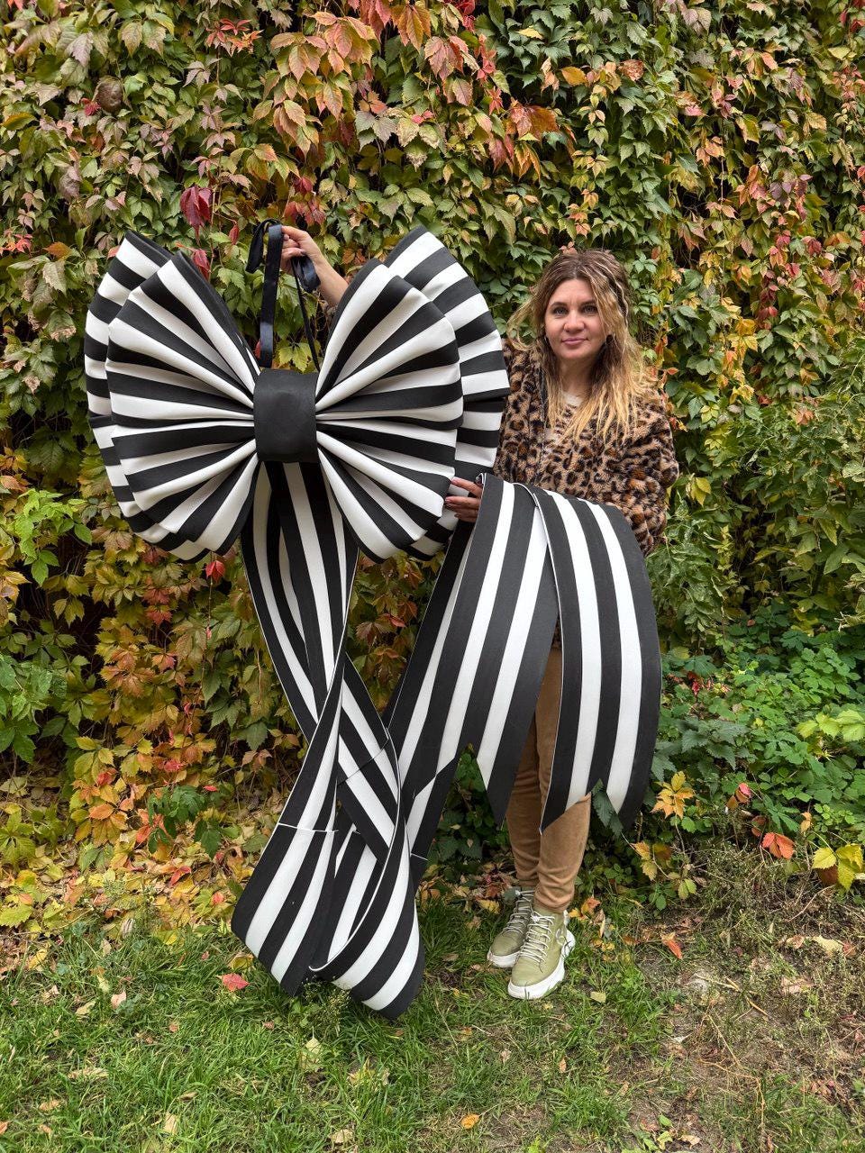 May include: A large, decorative bow with black and white stripes. The bow has a black center and long, striped tails. The bow is held up in front of a background of green and red foliage.