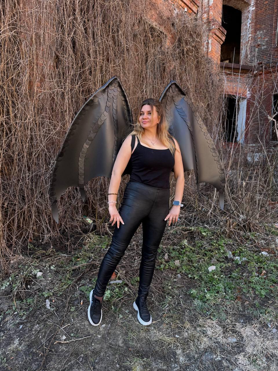 May include: A person wearing black faux leather pants, a black tank top, and black sneakers with white soles. The person is wearing large, black, dragon-like wings. The background features an old brick building and overgrown vegetation.