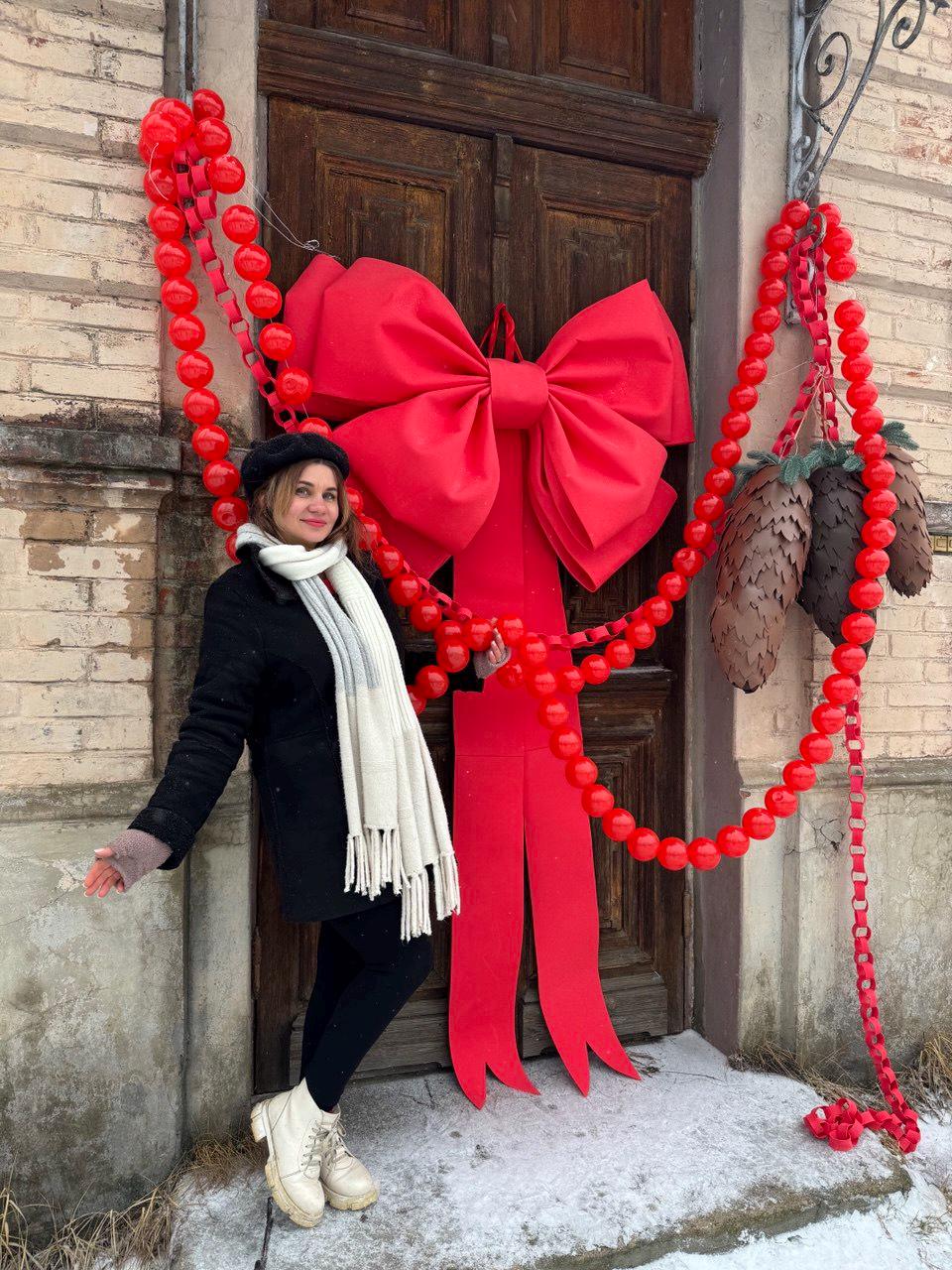 May include: A large red bow and matching garland adorn a wooden door. The garland is made of red spheres and paper chains. Two decorative pinecones hang from the garland. The door is set against a brick wall.