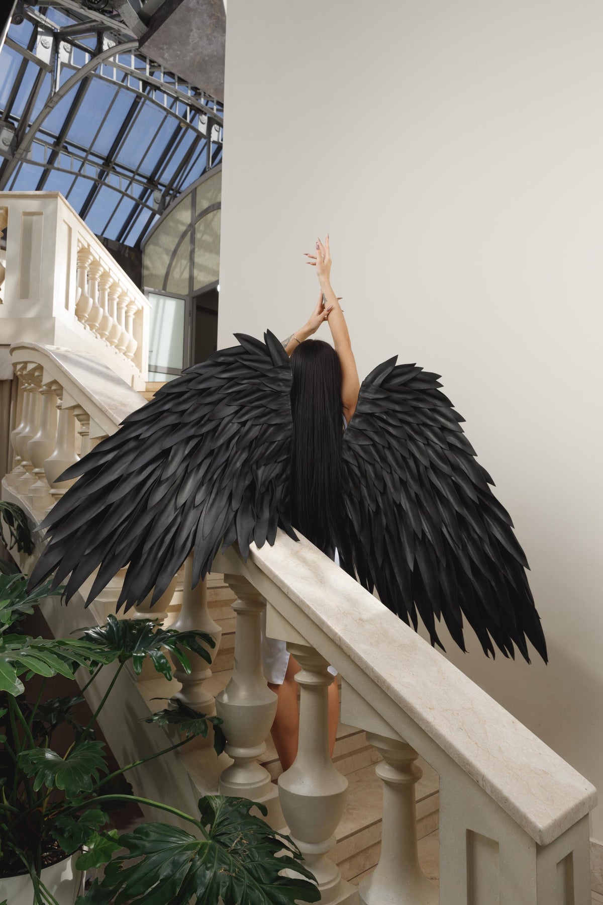 May include: A person with long black hair and large black feathered angel wings stands on a marble staircase. The wings are spread wide, and the person's arms are raised above their head. The background includes a glass ceiling and lush greenery.