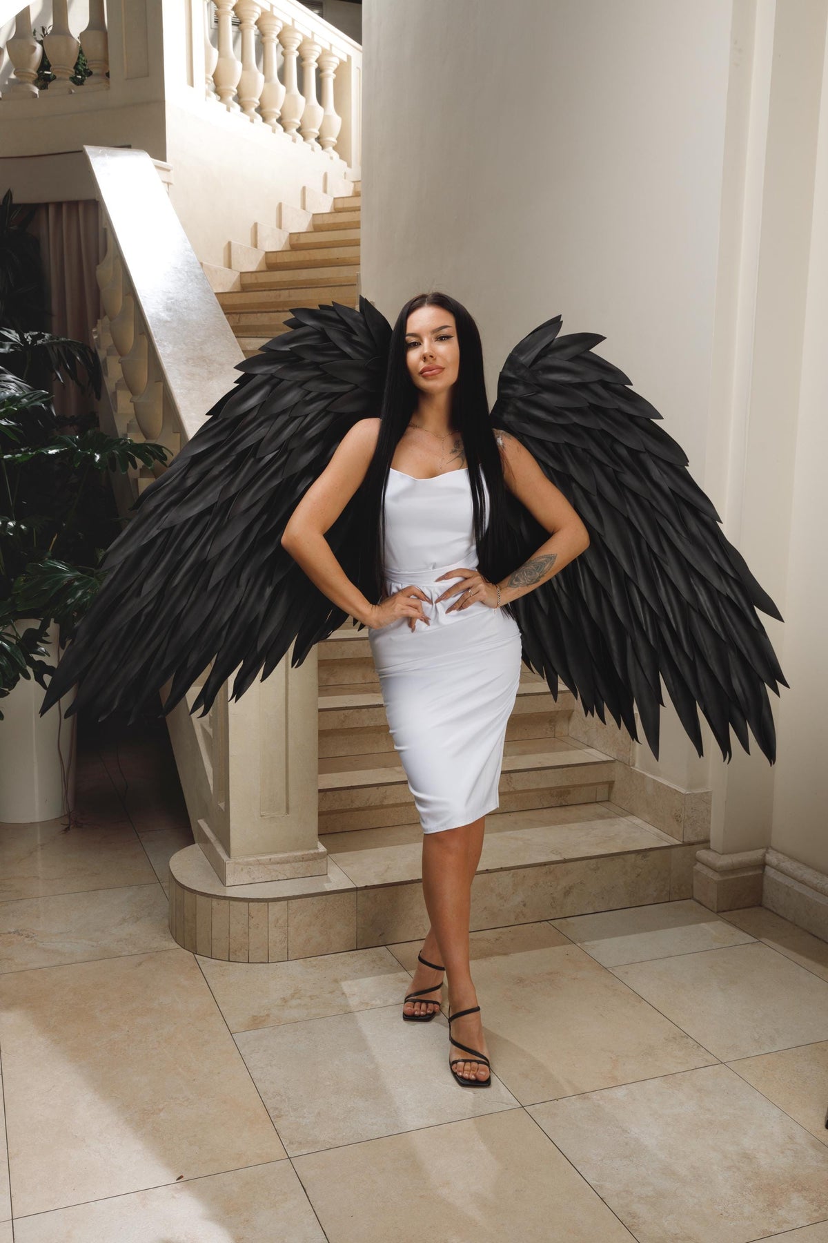 May include: A woman wearing a white dress and black sandals stands in front of a staircase, posing with large black feathered angel wings. The wings span wide, creating a dramatic contrast against the white dress and the neutral-toned background.