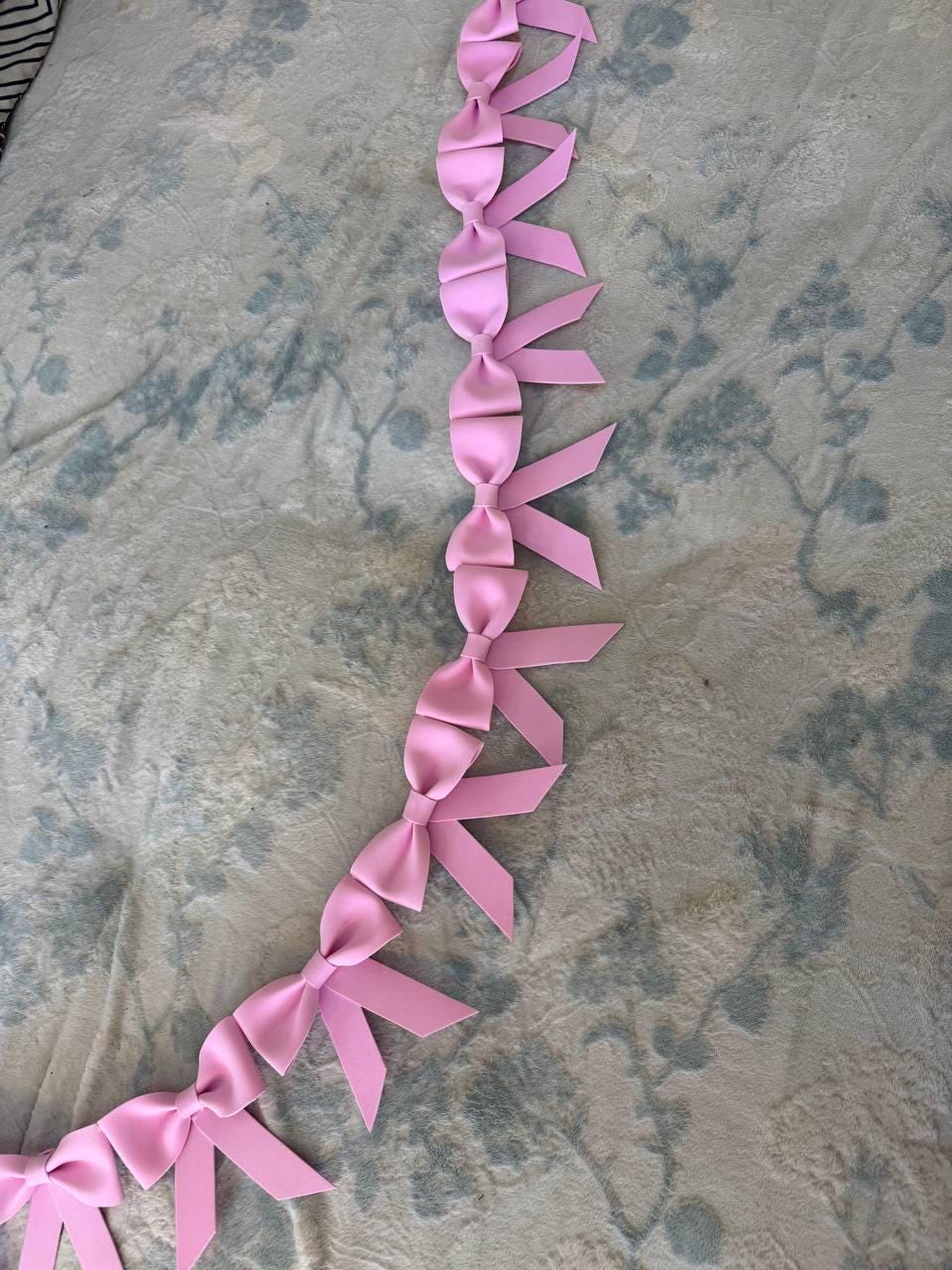 May include: A string of pink satin bows arranged in a vertical line. Each bow is tied and has long, pointed ribbon tails. The bows are a soft, pastel pink color and are set against a patterned background.