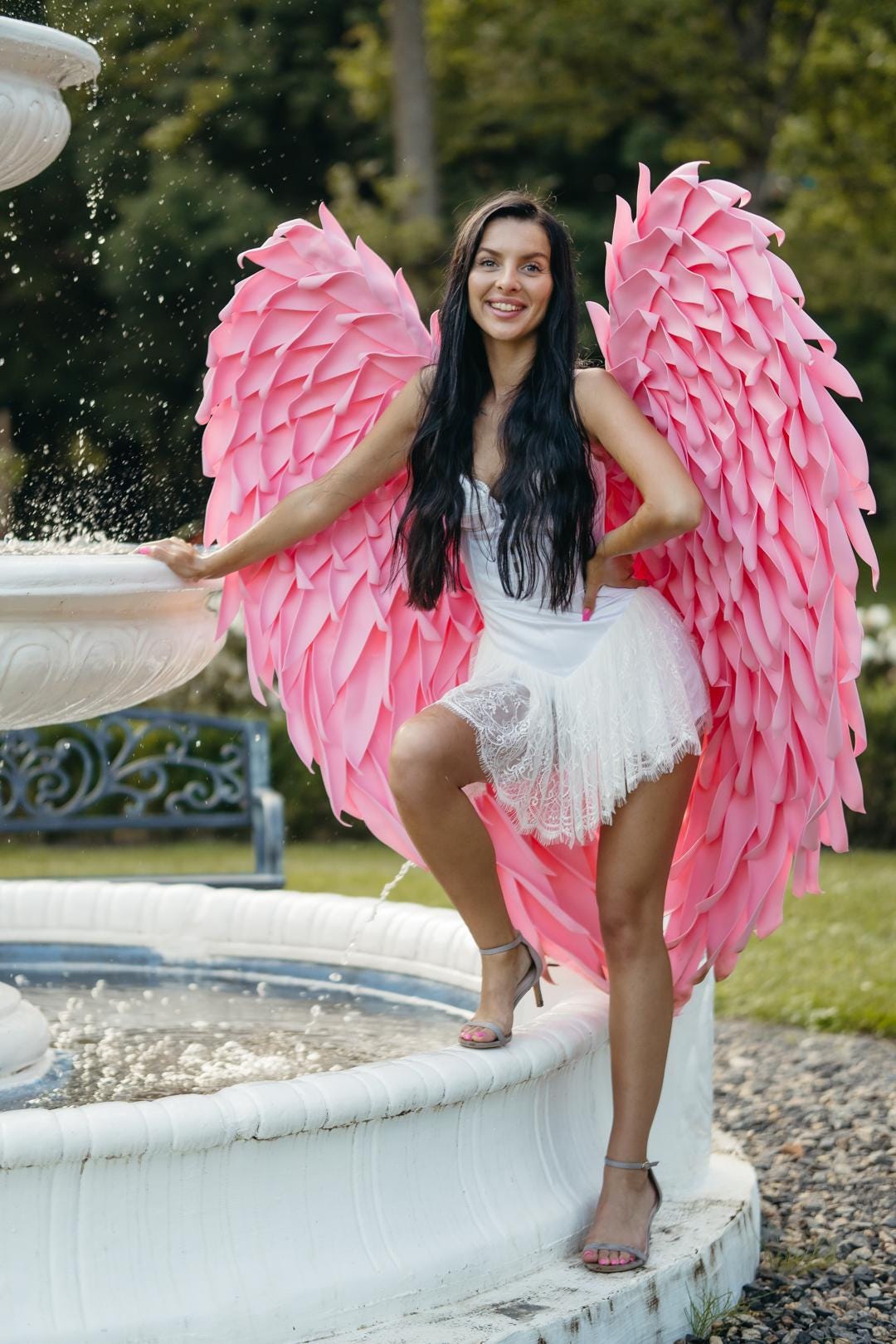 May include: A woman wearing a white dress and pink angel wings poses near a fountain. The large wings are a vibrant pink, and the dress has a lace trim. The background includes greenery and a decorative fountain.