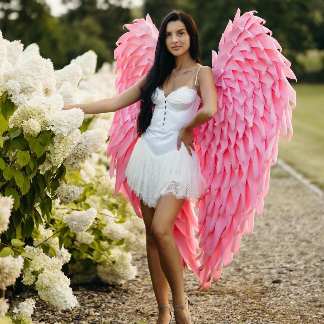 May include: A woman wearing a white dress with a lace skirt and pink angel wings stands near white flowers. The wings are large and feathered, with a gradient of pink shades. The dress has a corset-style bodice.