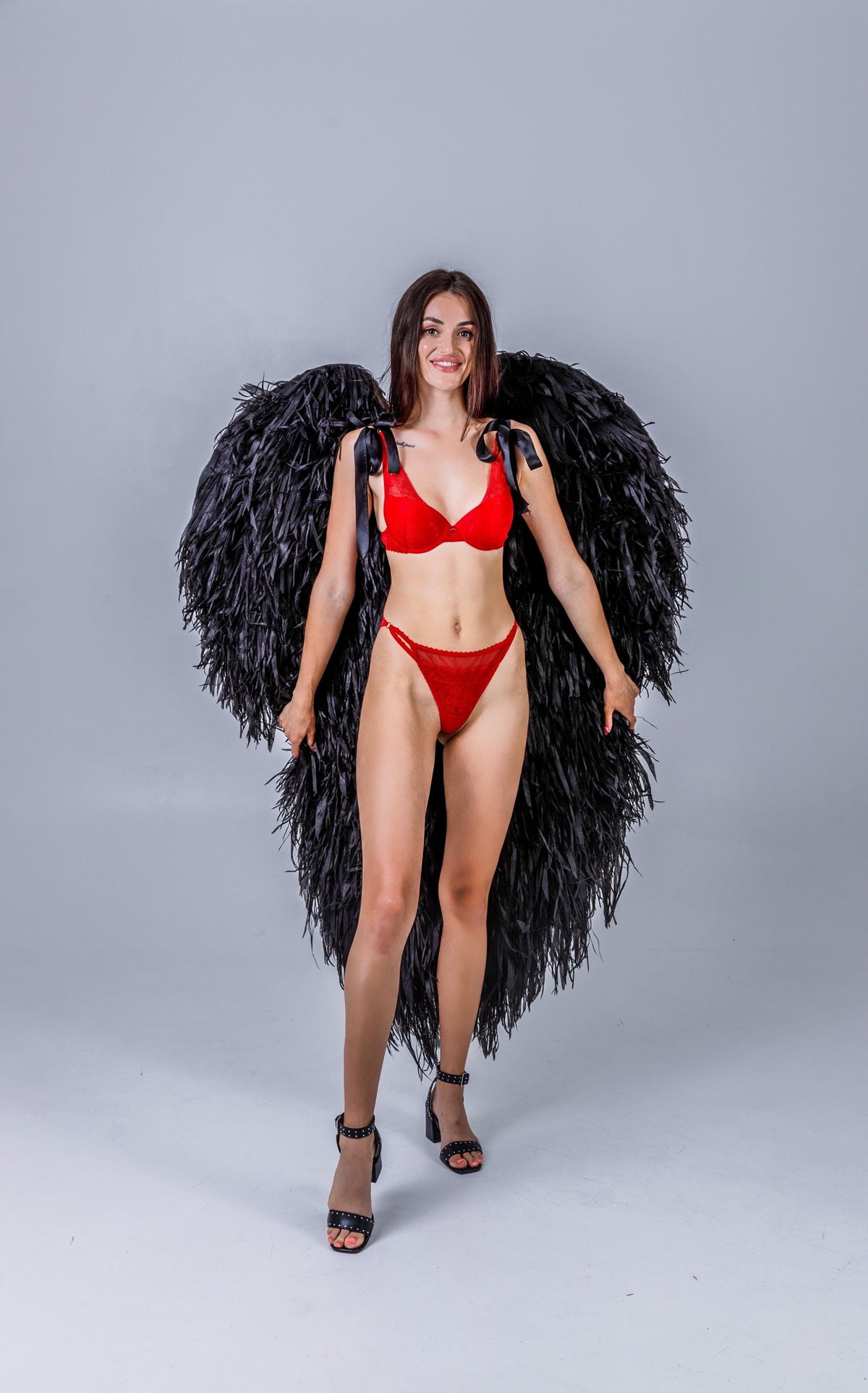 May include: A person wearing a red lingerie set and black feathered angel wings. The lingerie includes a bra with ribbon straps and a matching thong. The wings are large and black, and the person is wearing black high heels.