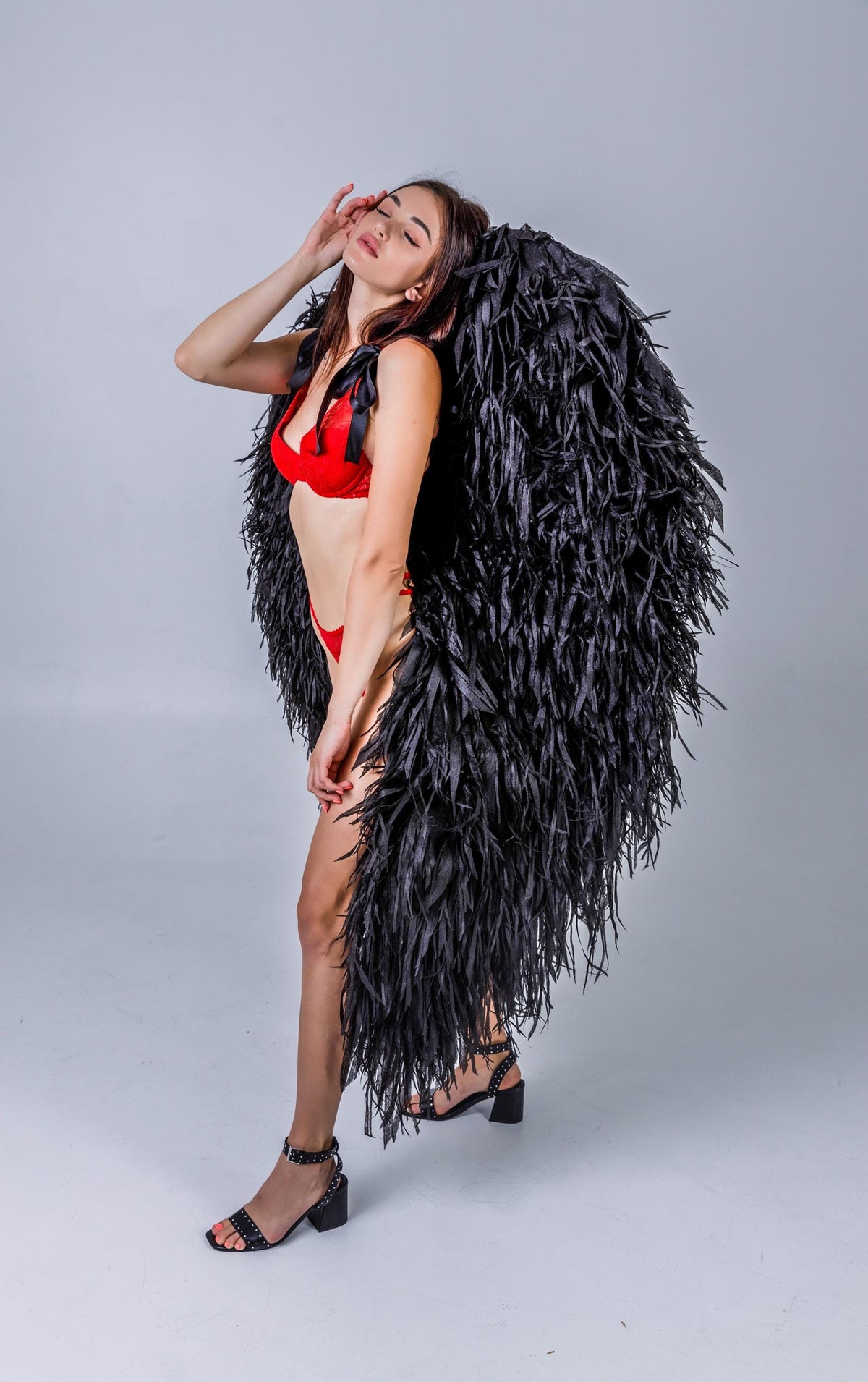 May include: A woman wearing a red lingerie set and black feathered angel wings. The wings are large and cover the back. She is also wearing black heeled sandals with studded details.