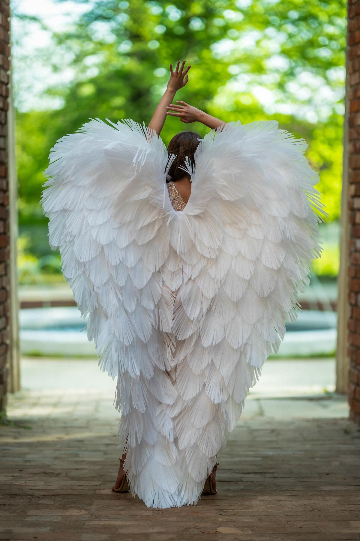 May include: A white feathered costume with large, layered wings. The costume is worn by a person with their arms raised. The background includes a brick structure and greenery, creating a dramatic and artistic effect.