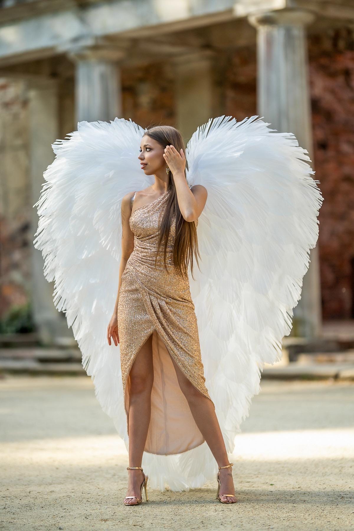 May include: A woman wearing a gold sequined one-shoulder dress and large white feathered angel wings. The dress has a high slit, and the woman is wearing gold heels. The background features a stone building with columns.