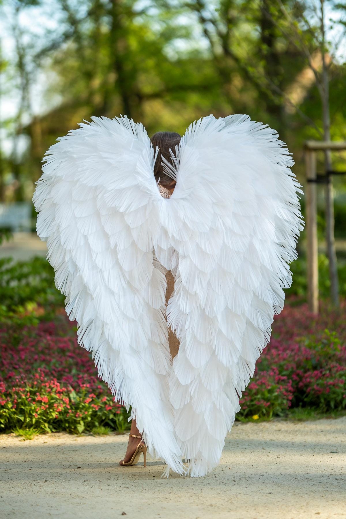 May include: A person wearing large, white feather angel wings. The wings are heart-shaped and cover the person's back. The person is wearing gold high heels. The background is a park with trees and flowers.