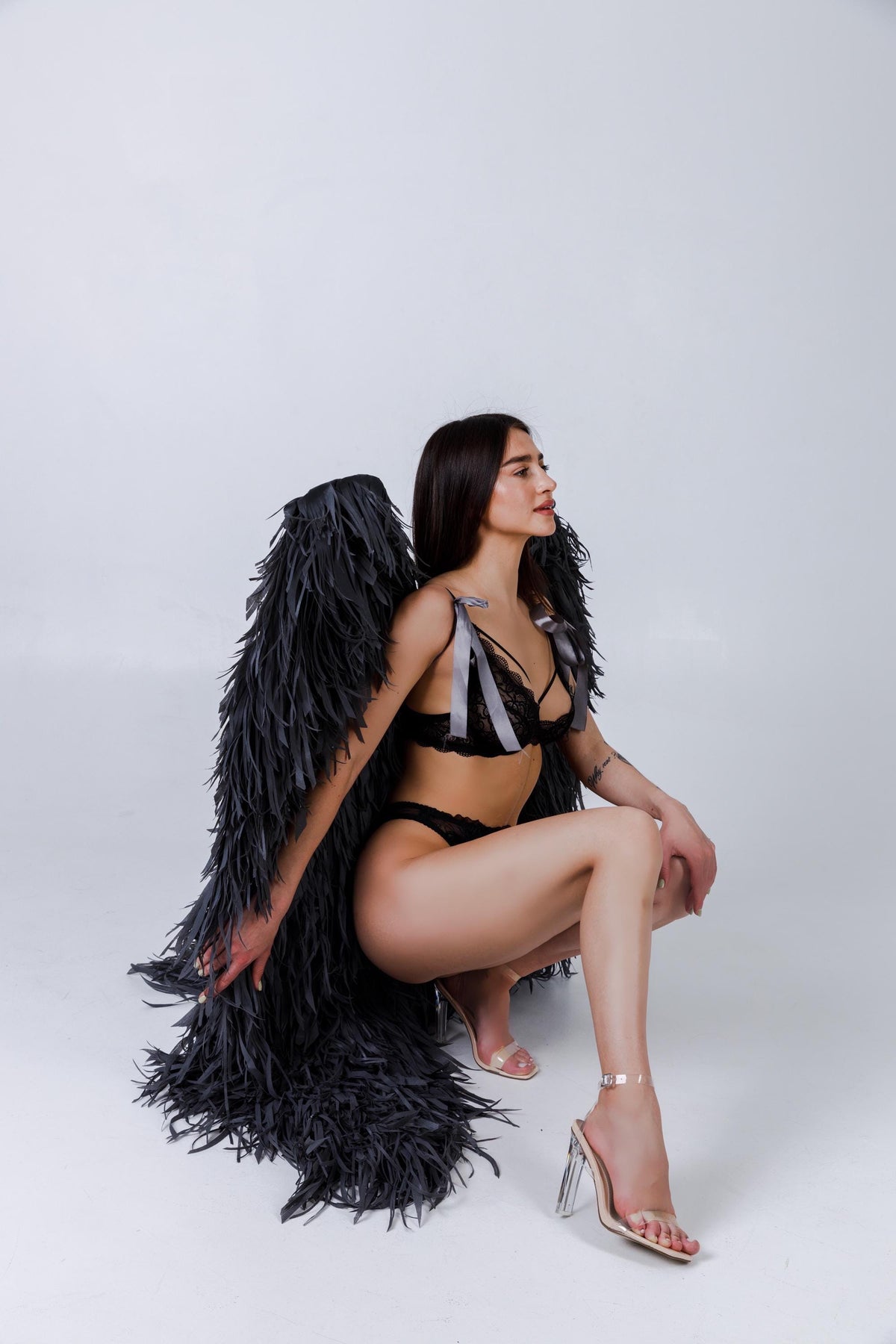 May include: Black lingerie set with matching feathery angel wings. The lingerie features lace detailing and ribbon accents. The wings are large and dark, creating a dramatic effect. Clear heeled sandals complete the look.