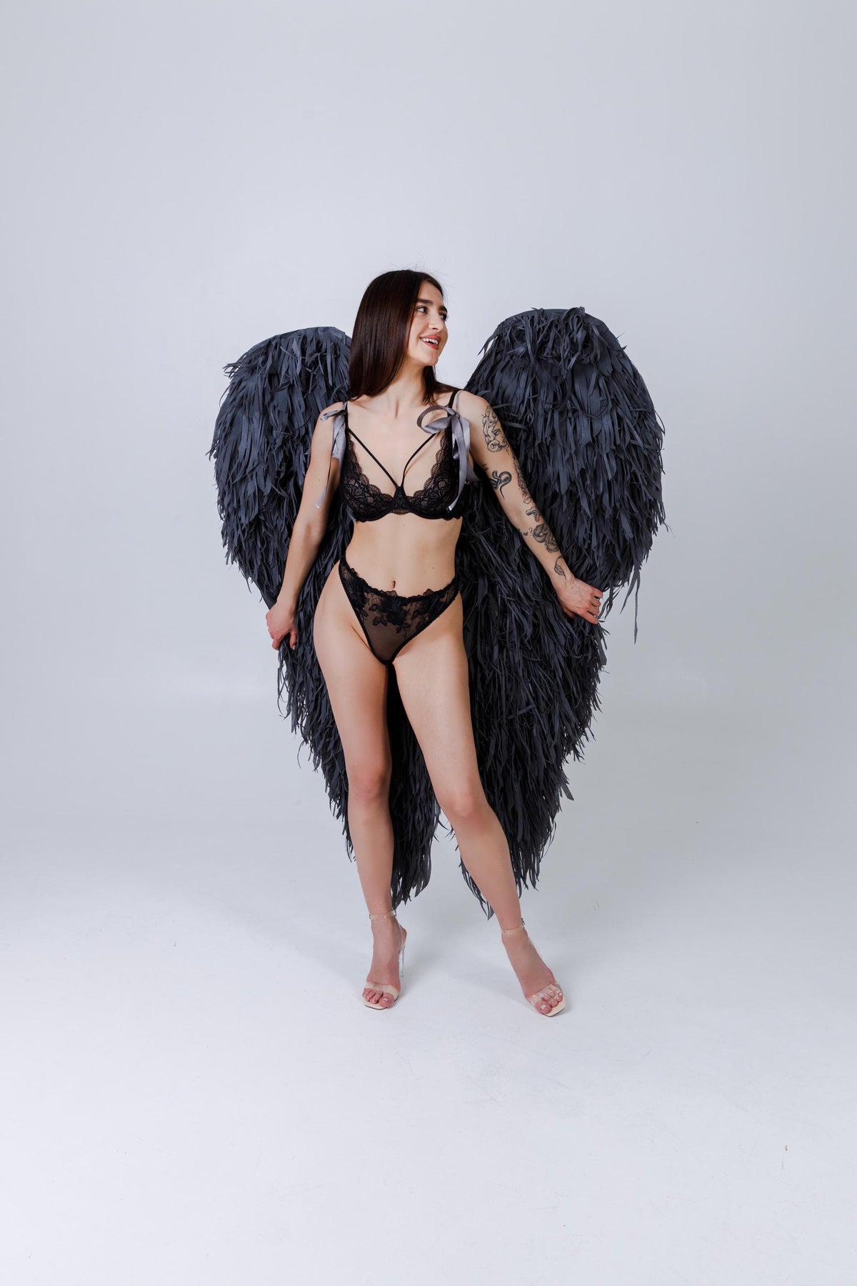 May include: Black lingerie set with a bra, high-waisted bottoms, and large black feathered wings. The bra has thin straps and the bottoms have floral lace details. The wings are large and dark, creating a dramatic effect.