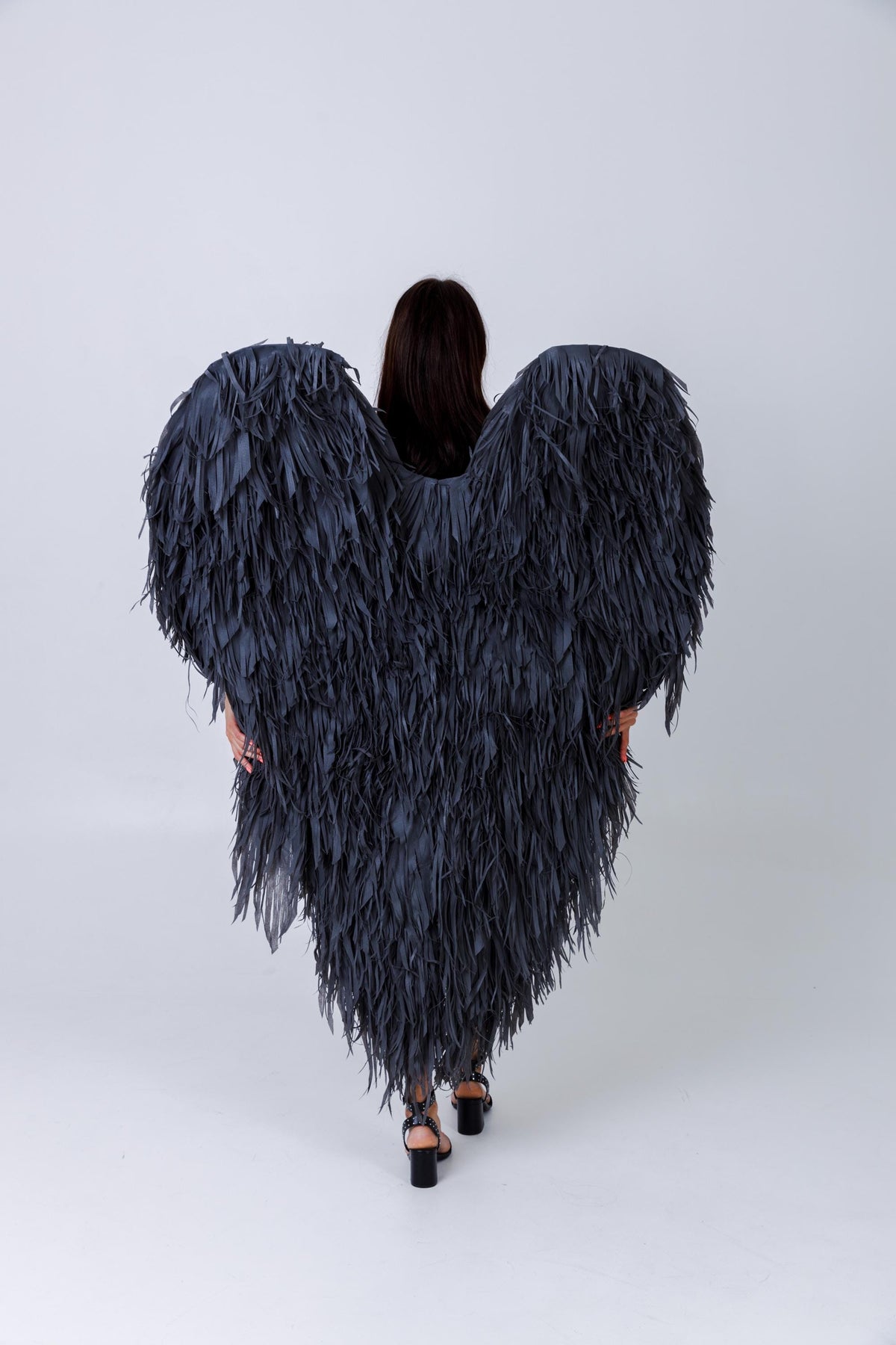 May include: A person wearing large, dark gray feather wings. The wings are heart-shaped and cover the person's back. The feathers are long and shaggy, creating a dramatic effect. The person is wearing black heels.