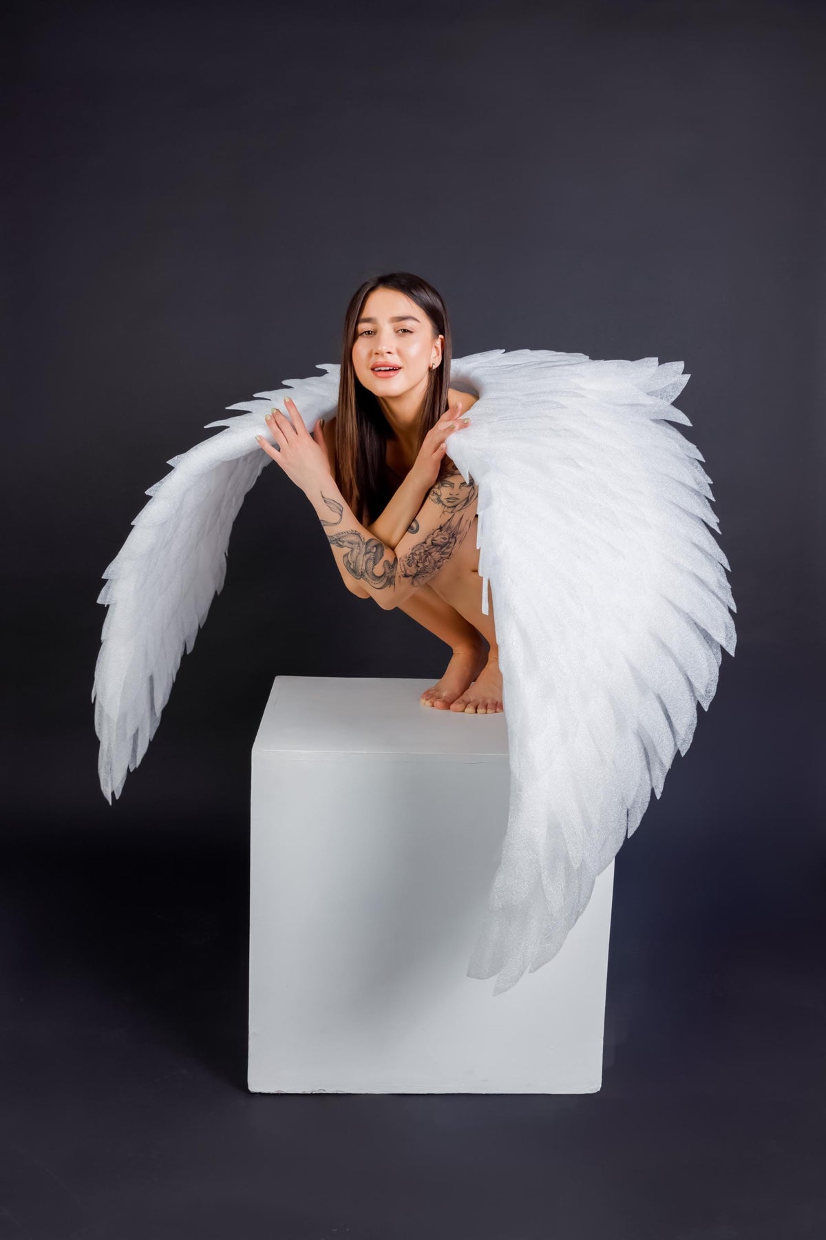 May include: A person wearing large, white angel wings, posing on a white cube against a dark gray background. The wings are detailed with layered feathers. The person has tattoos on their arms and is looking at the camera.