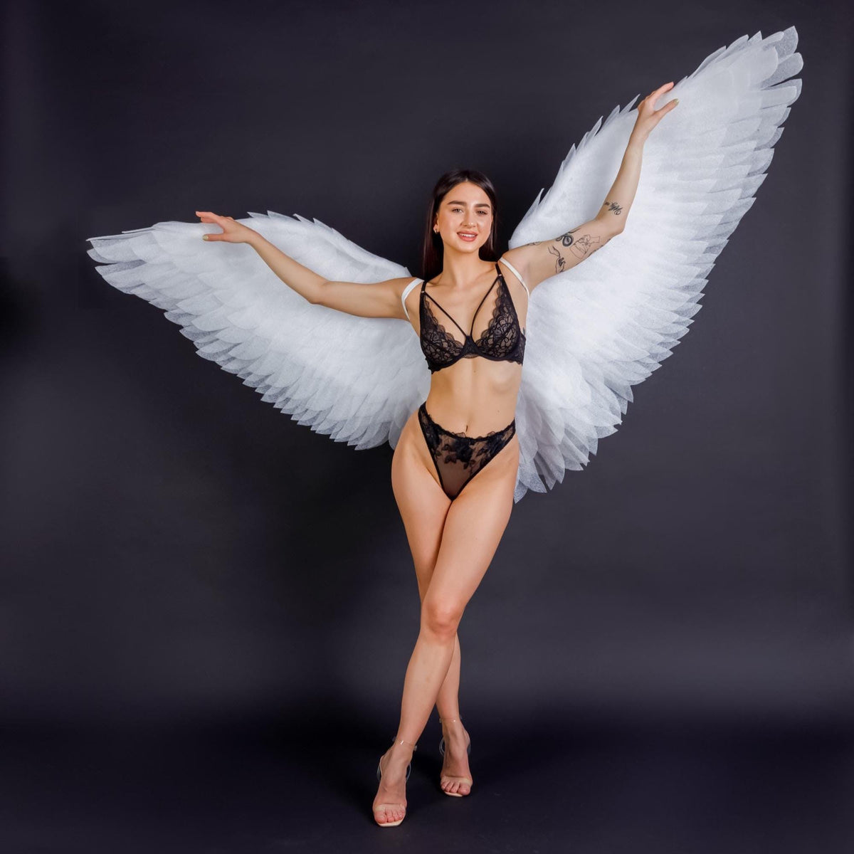 May include: A woman wearing a black lace lingerie set and large white feathered angel wings against a dark background. The lingerie includes a bra and high-waisted bottoms. The wings are spread wide, creating a dramatic pose.