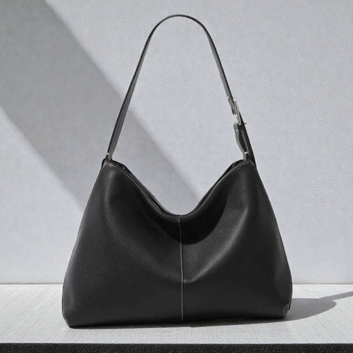 May include: A black leather shoulder bag with a single strap and silver-tone hardware. The bag has a curved shape and a white stitched seam down the center. The bag is sitting on a white surface.