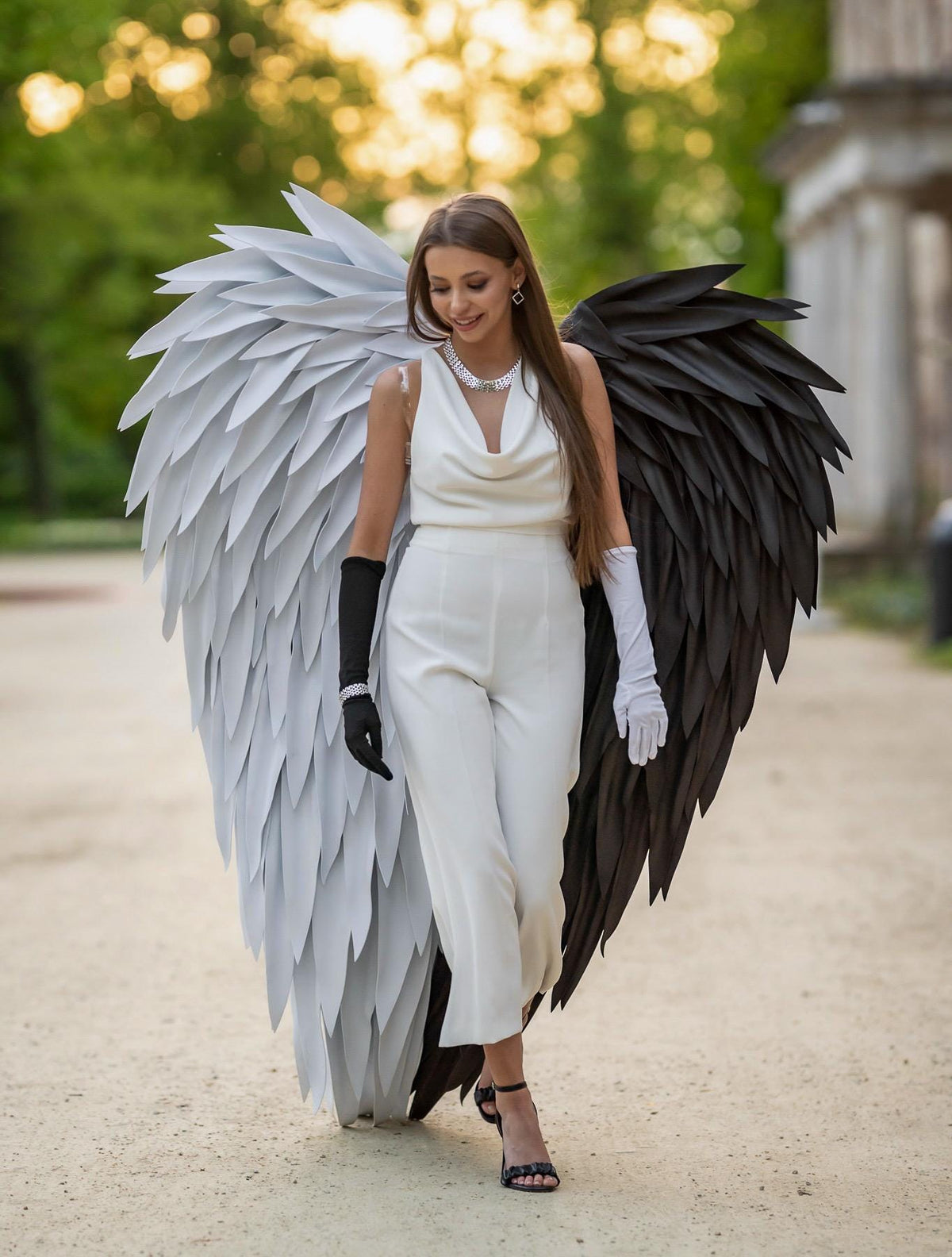 May include: A white sleeveless jumpsuit with a cowl neckline paired with large, oversized wings. The left wing is white and the right wing is black. The outfit includes one black glove on the right hand and one white glove on the left hand.