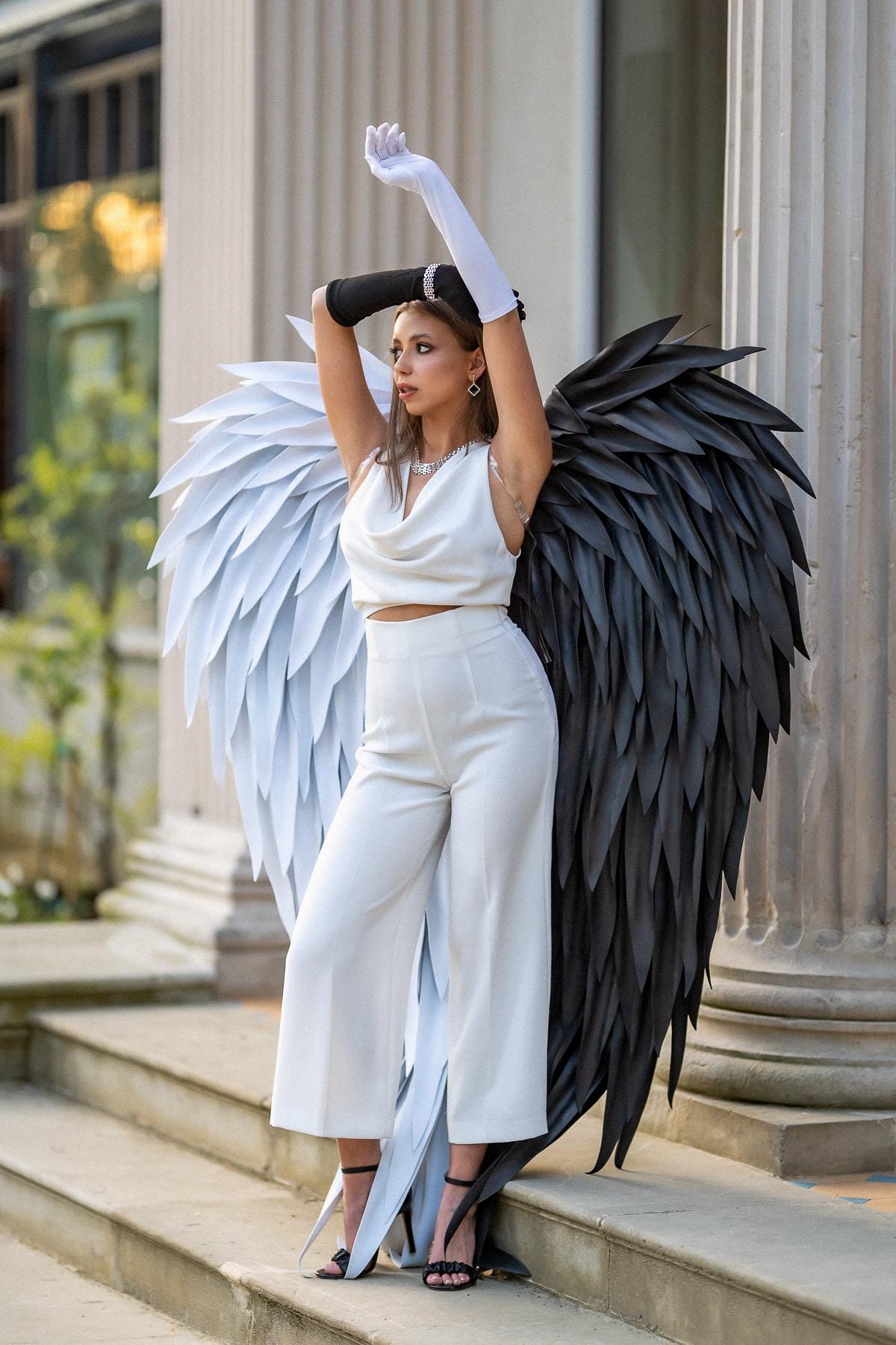 May include: A white sleeveless cowl-neck crop top and matching high-waisted wide-leg trousers paired with large decorative wings, one side white and one side black. The outfit includes one long white glove and one long black glove with black sandals.