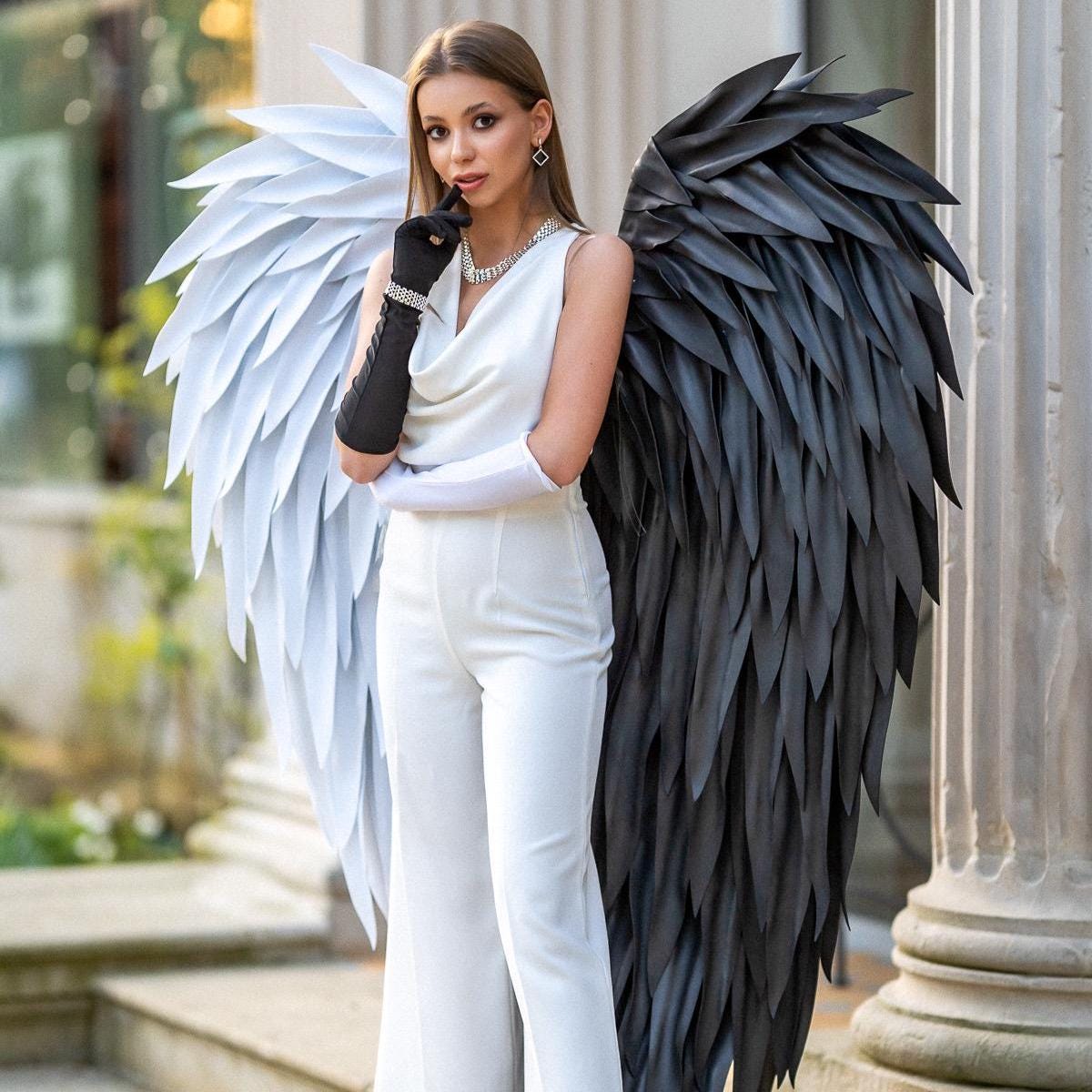May include: A white sleeveless jumpsuit with a cowl neckline paired with a pair of large decorative wings. The left wing is white and the right wing is black, both featuring layered feather-like segments. One black glove and one white glove are worn.