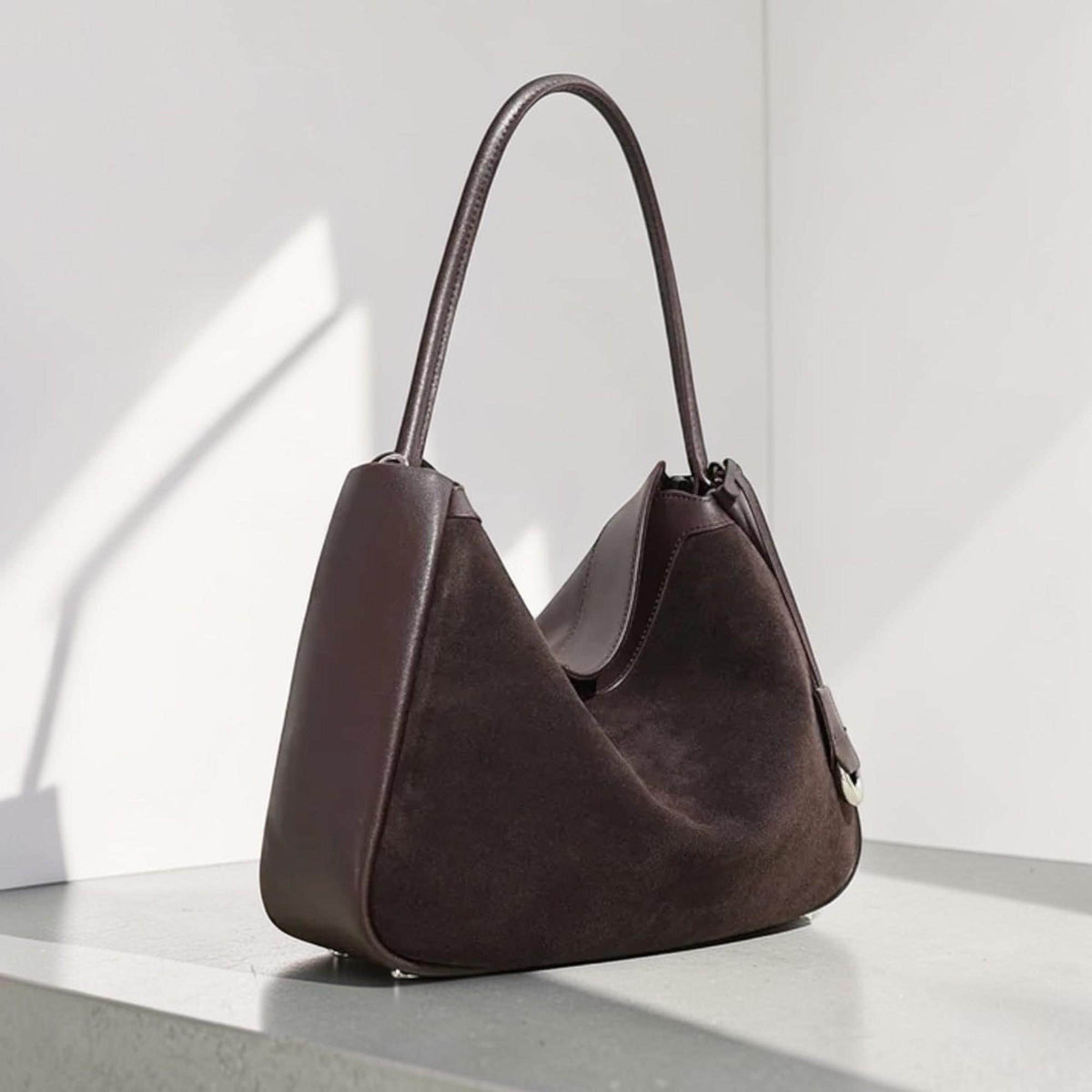 May include: A dark brown suede shoulder bag with a curved handle. The bag has a smooth leather trim and a structured design. The bag is set against a neutral background, highlighting its shape and texture.