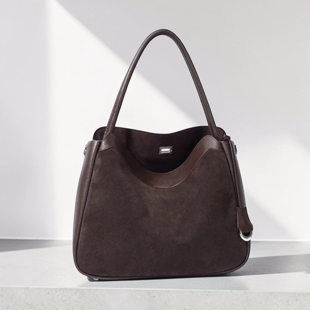 May include: A brown leather tote bag with a suede-like texture. The bag has a curved top edge, a long handle, and a smaller handle with a silver ring. The bag is sitting on a white surface.