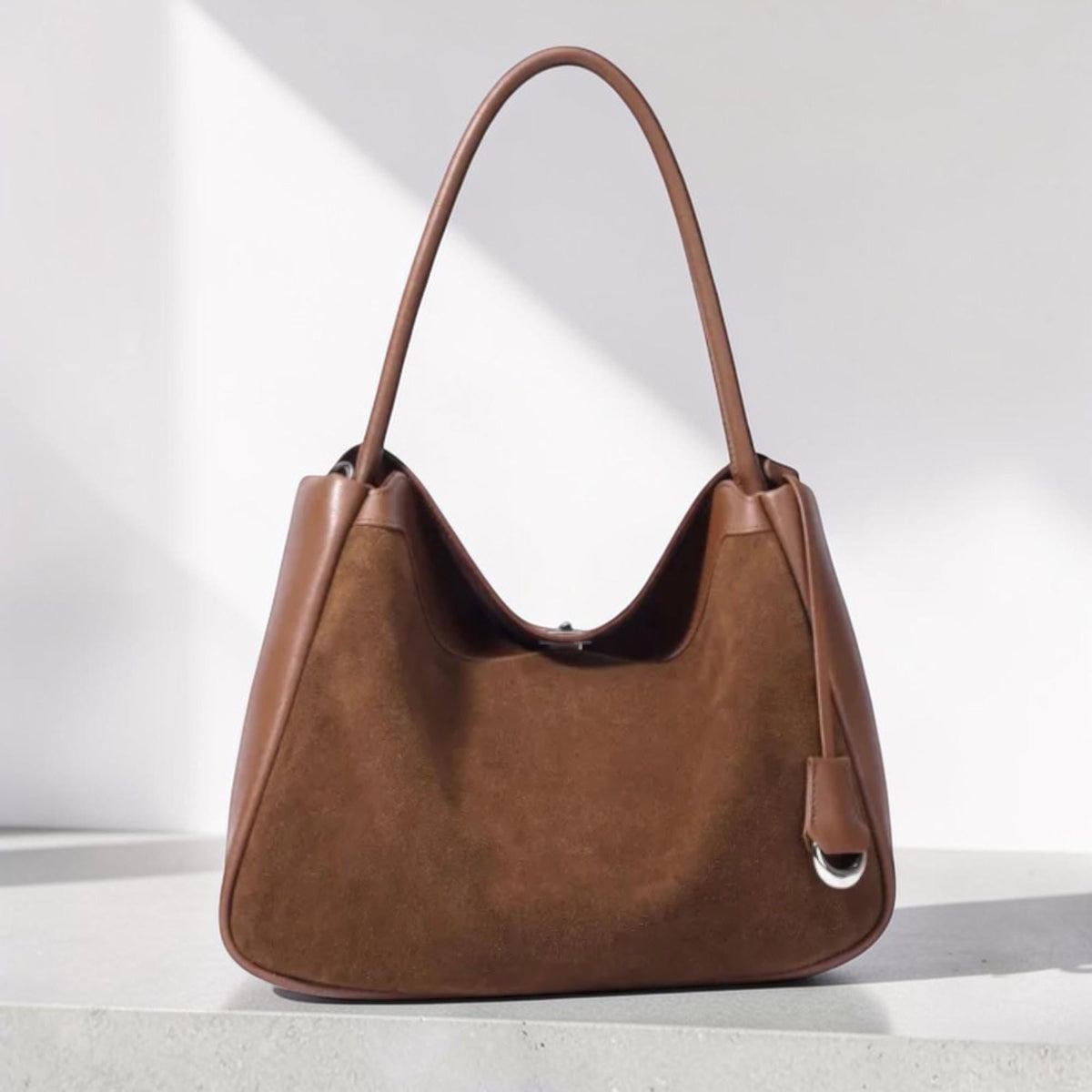 May include: A brown suede shoulder bag with a curved top and a single handle. The bag has a leather trim and a small leather tag with a silver-colored ring. The bag is sitting on a white surface.