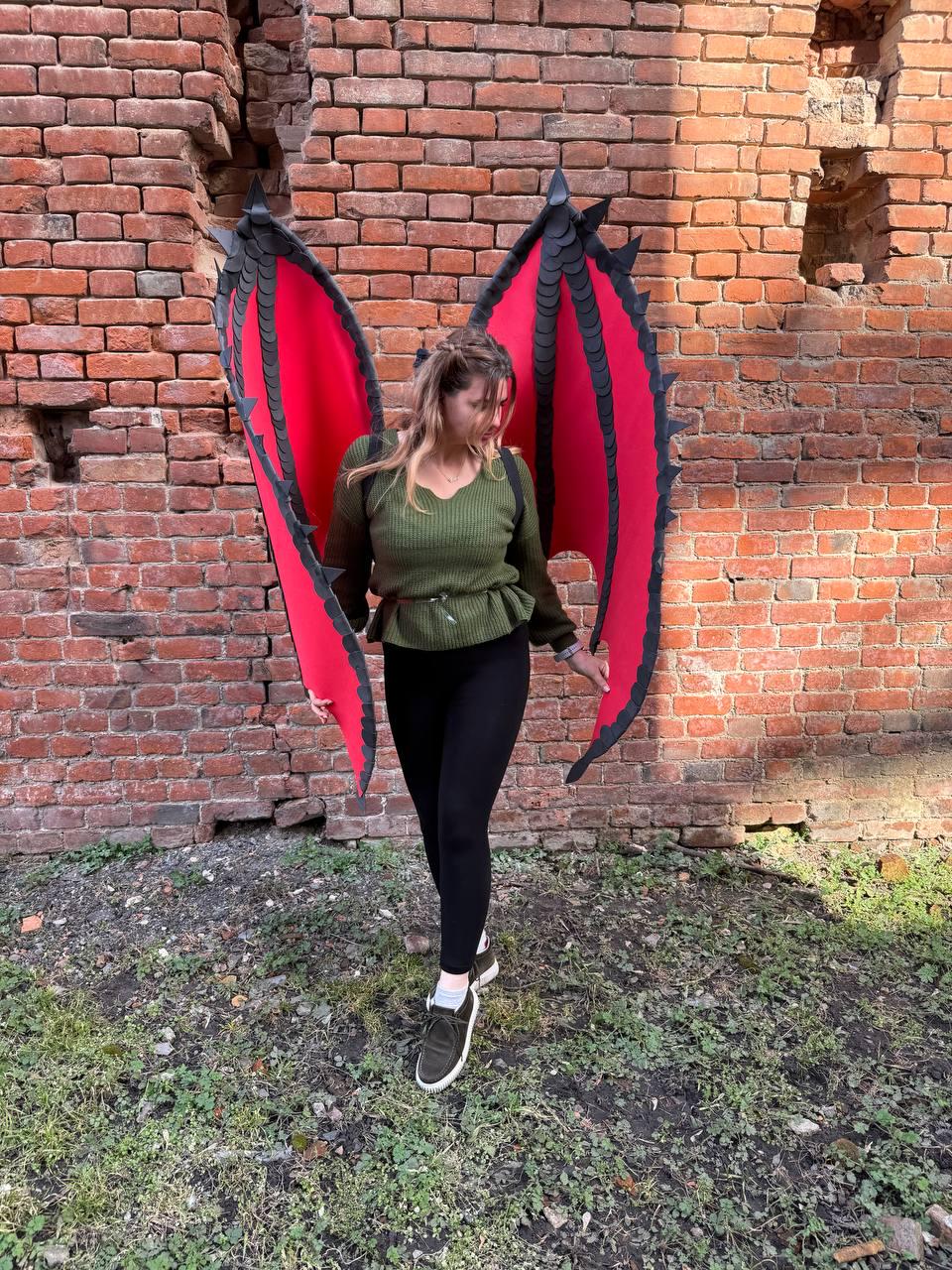 May include: Red and black dragon wings with a detailed, scaled design. The wings are spread wide, showcasing the vibrant red interior and black edging. The wings are worn by a person in a green top and black leggings, standing in front of a brick wall.