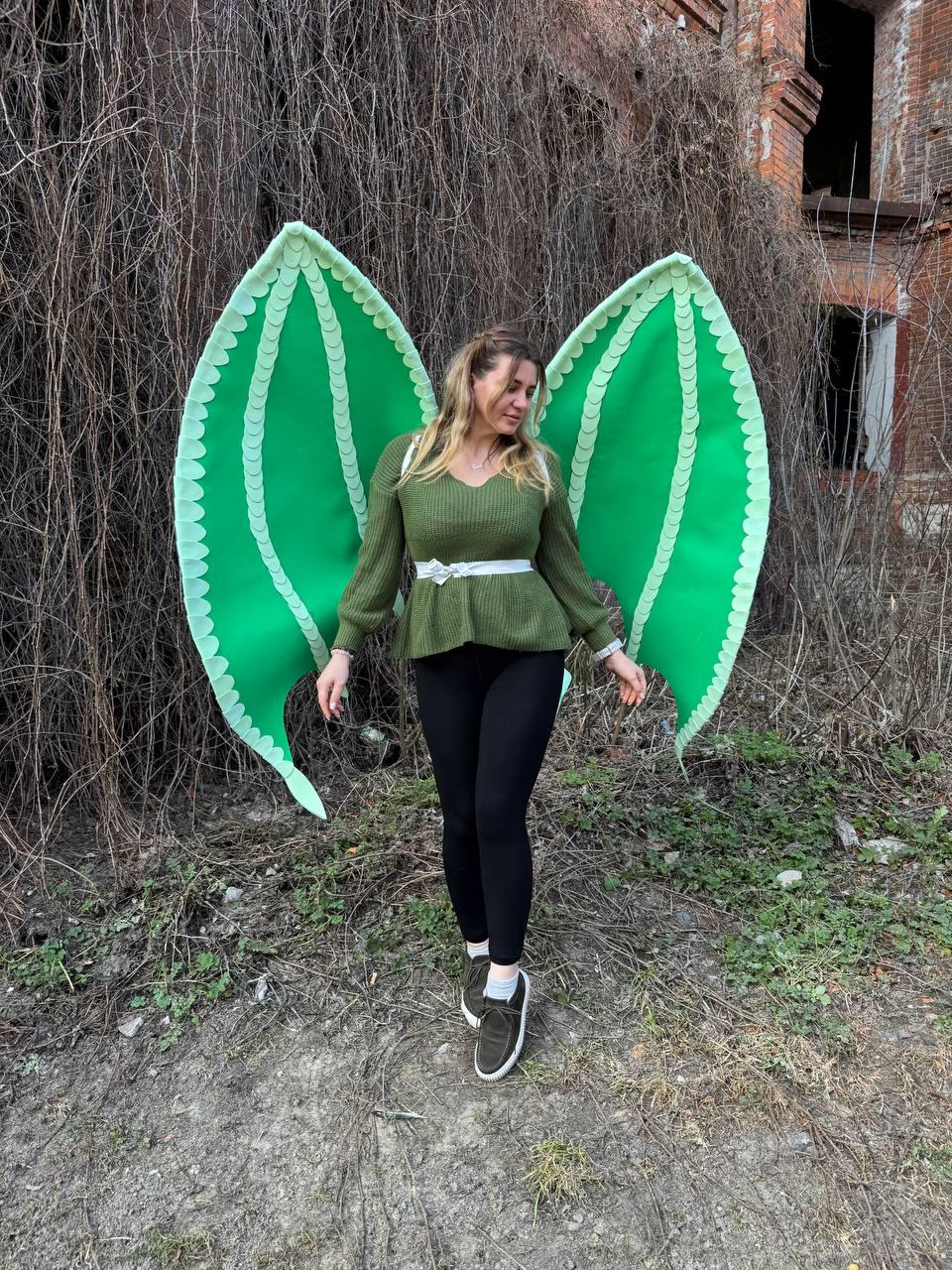 May include: Large, bright green dragon wings with scalloped edges. The wings are worn by a person wearing a green sweater, white belt, and black leggings. The wings are the focal point, with a backdrop of overgrown foliage and a weathered building.